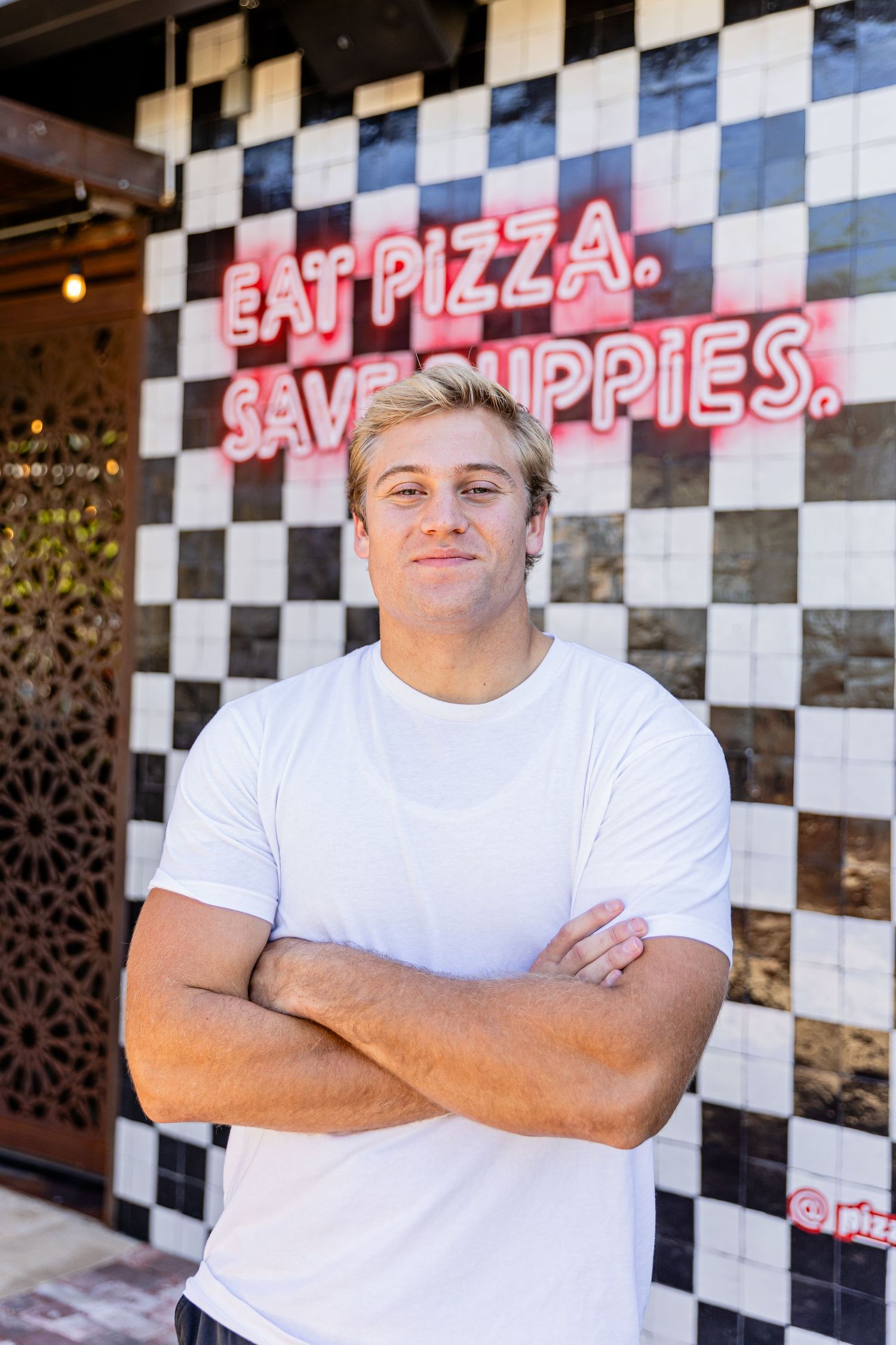 Young man in white t-shirt stands arms crossed in front of checkered wall with Eat Pizza Save Puppies neon sign.