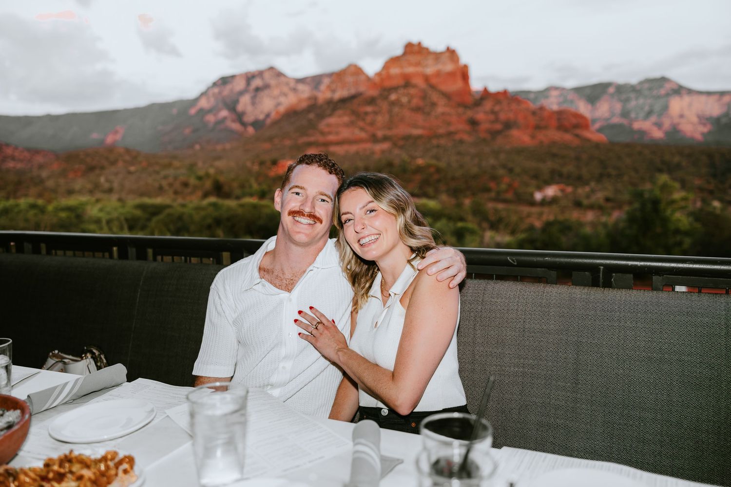 Couple smiling together at outdoor dining table with red rock Sedona mountains in background at dusk.