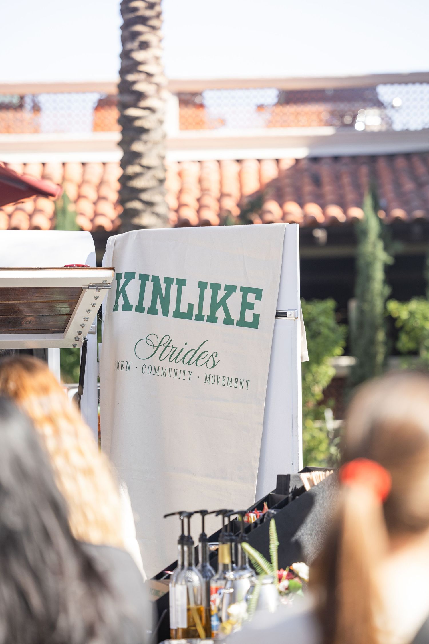 A banner reading Kinlike Strides Women Community Movement hangs at an outdoor event near a tiled roof building.