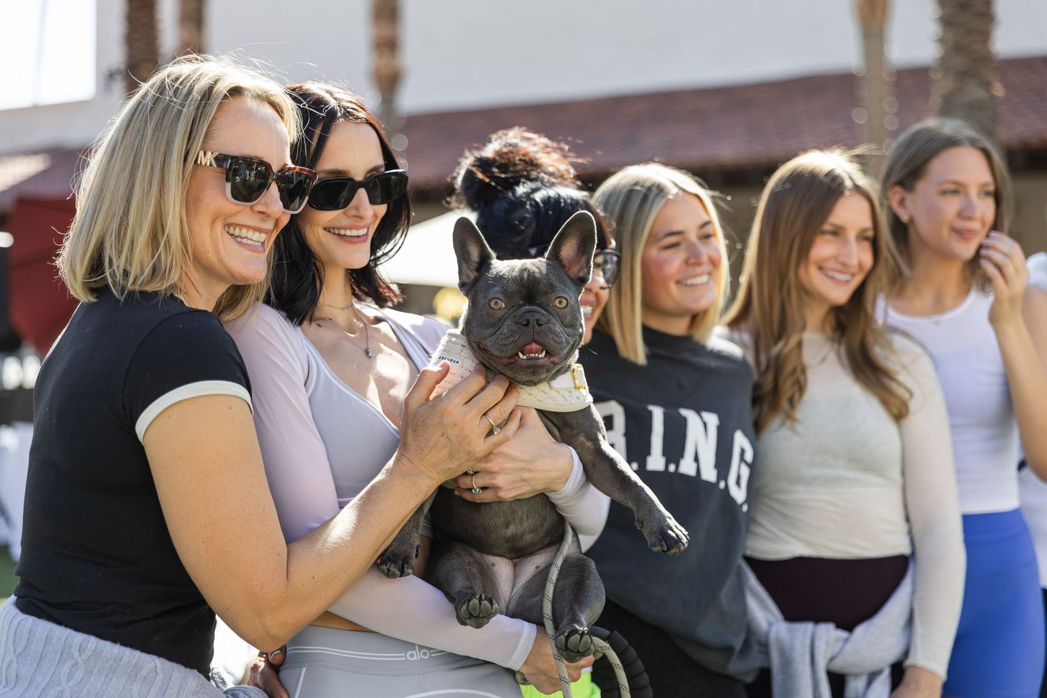 Group of women smiling outdoors holding a French Bulldog at a social event.