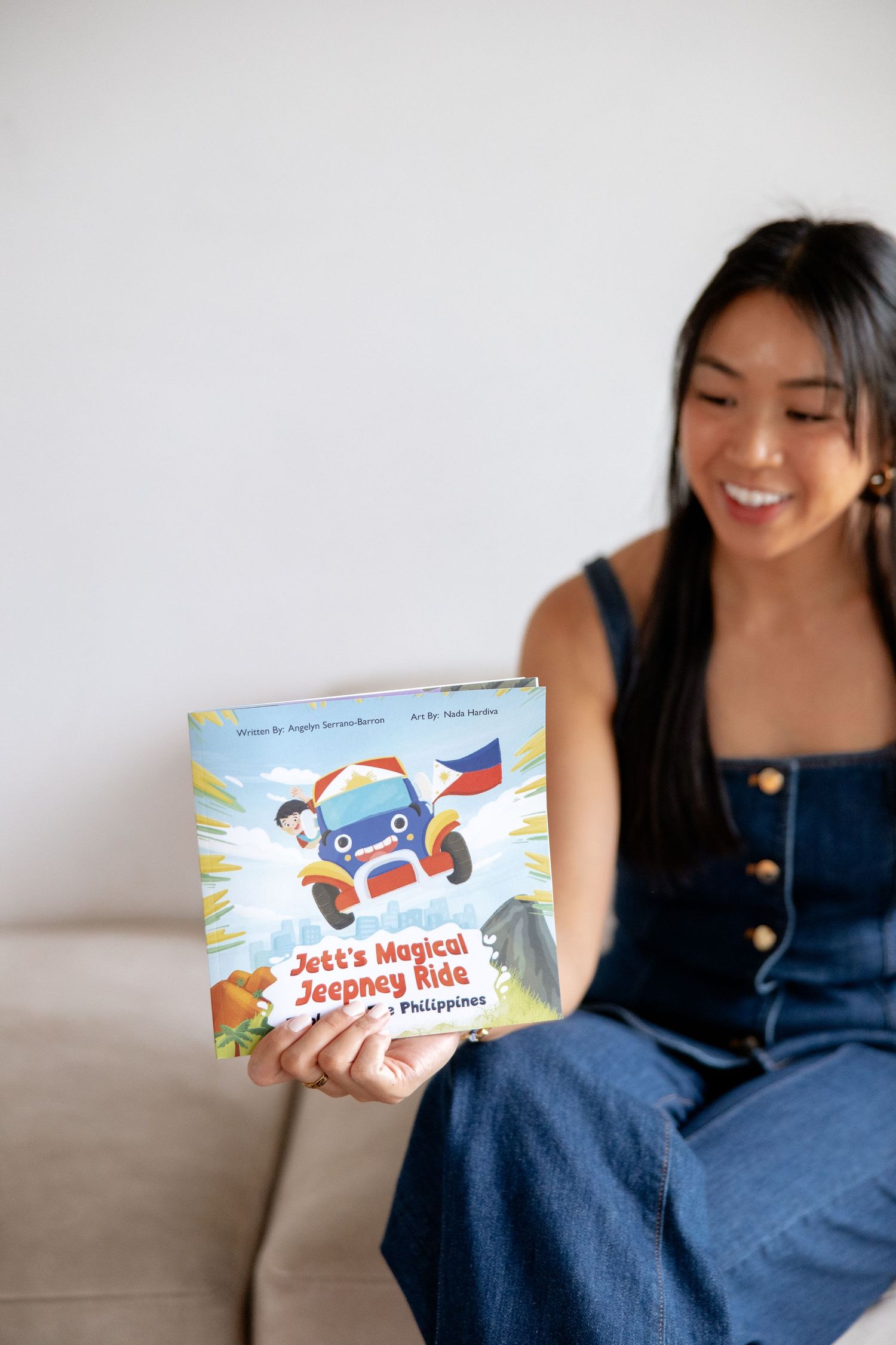 Smiling woman holding Jett's Magical Jeepney Ride children's book about the Philippines while sitting on a couch.