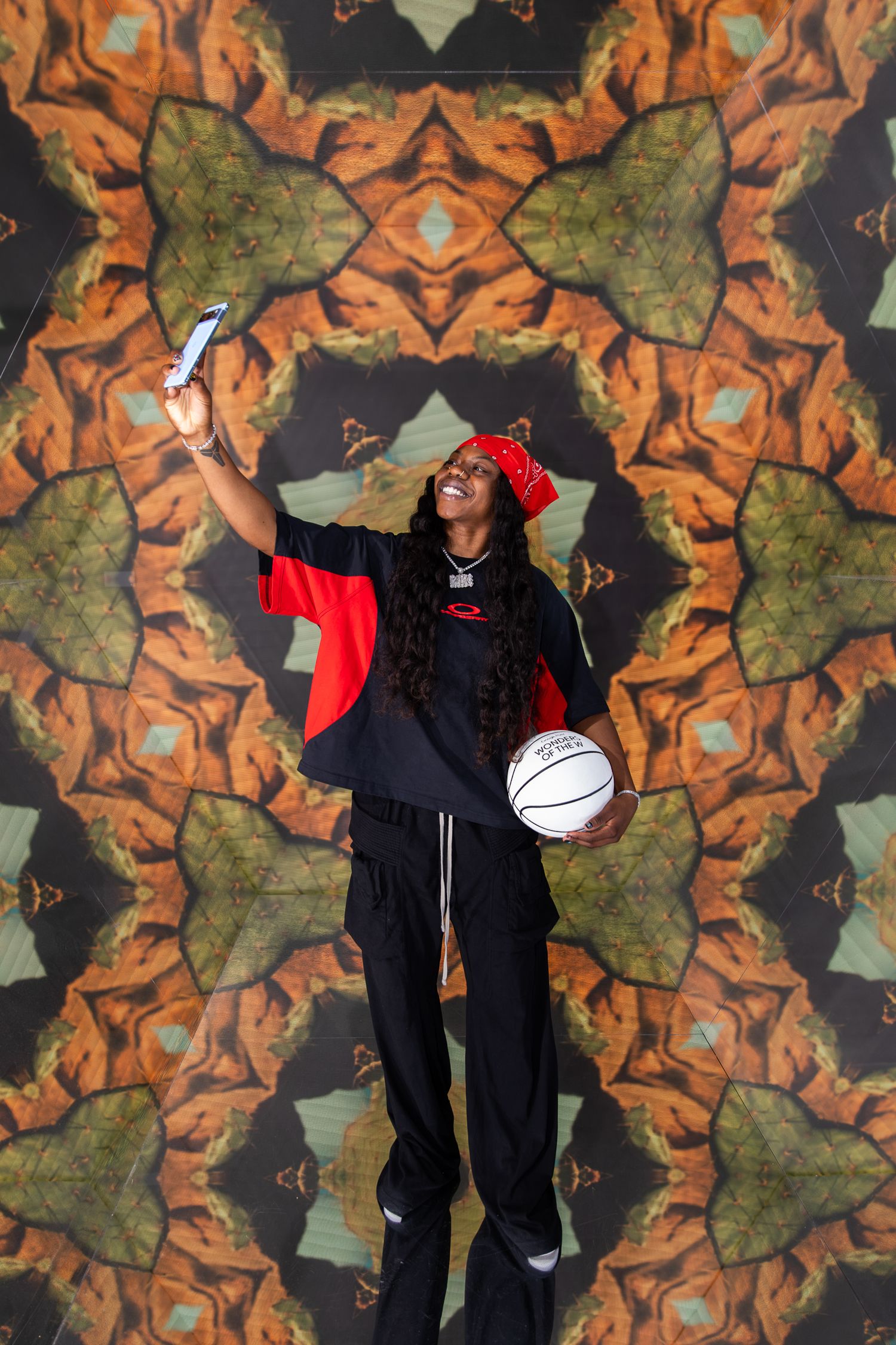 Smiling woman in red cap and black outfit taking selfie while holding basketball against colorful kaleidoscope backdrop.