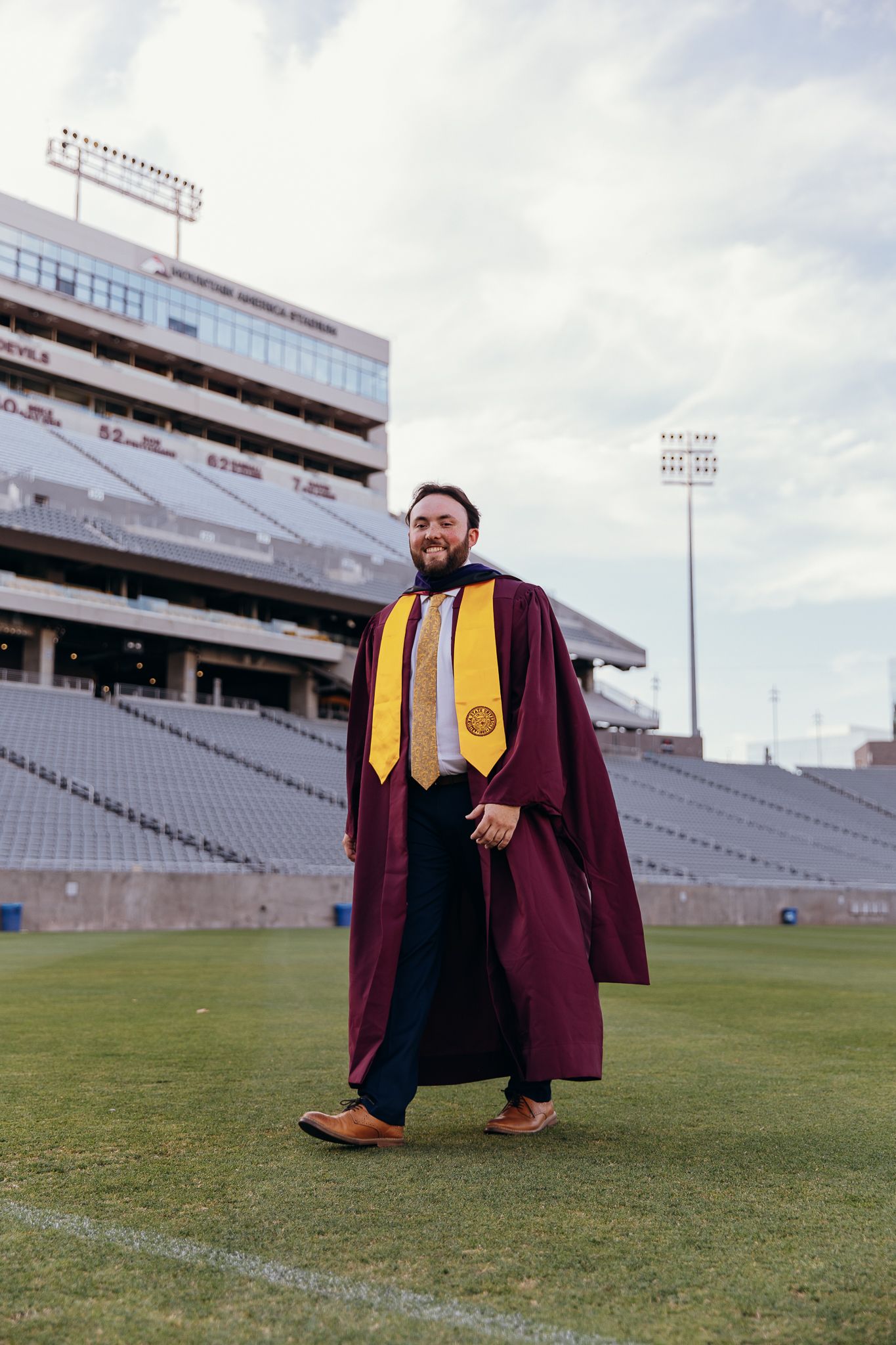 Graduate in maroon and gold ASU regalia standing on football field stadium.