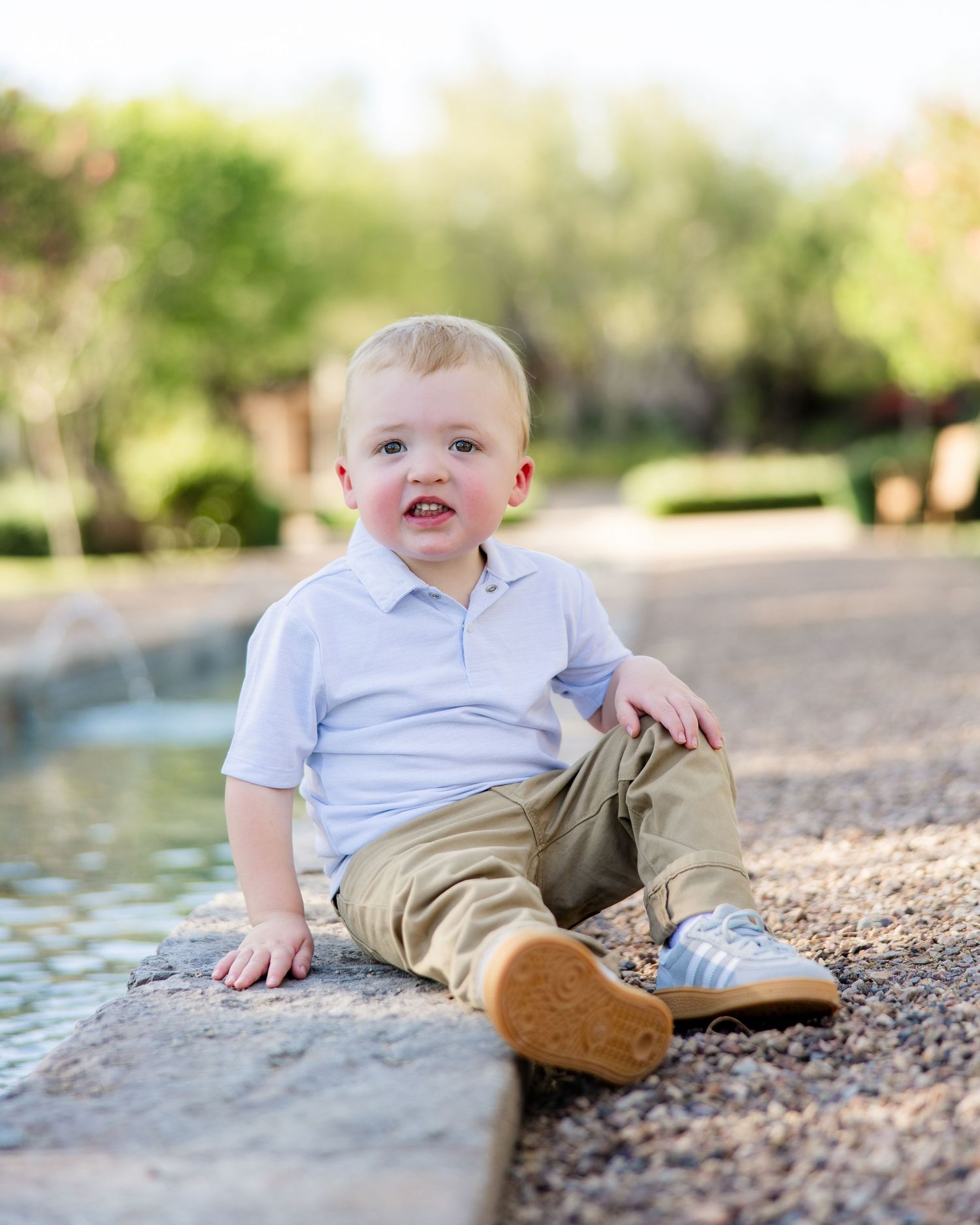 Happy blonde toddler boy in white polo sitting by fountain in sunny outdoor park.