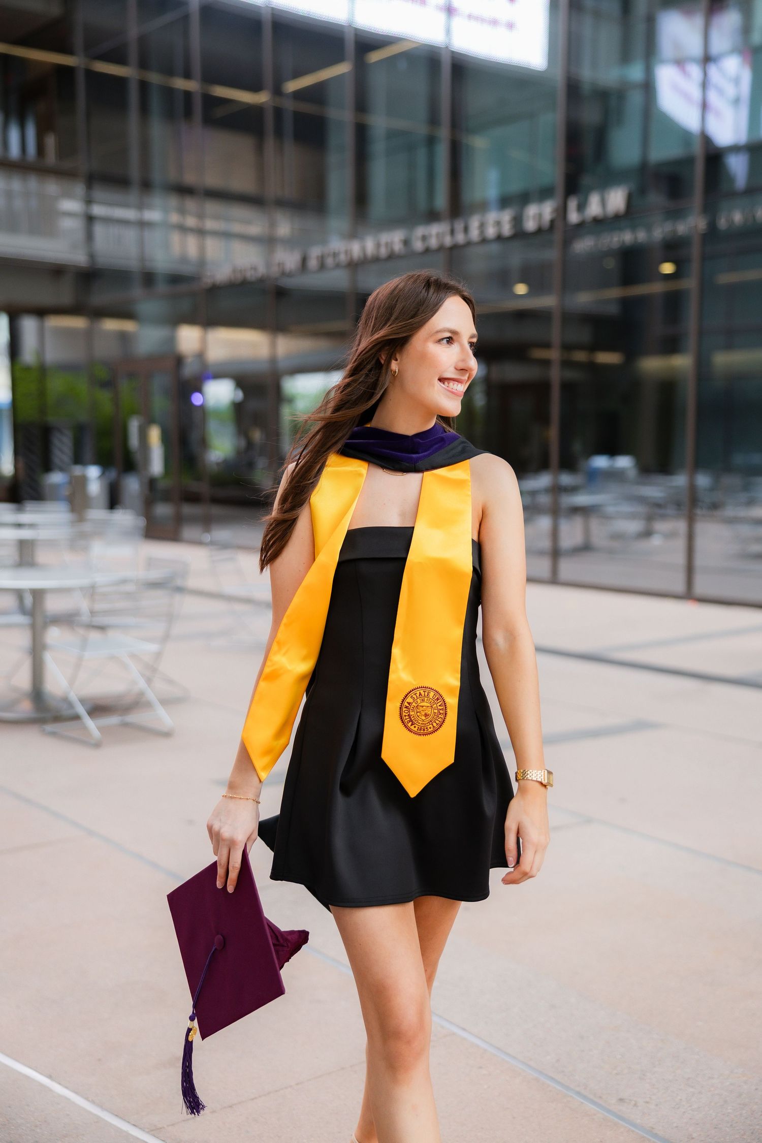 Brunette woman walks confidently in black dress with gold graduation stole outside law school building.
