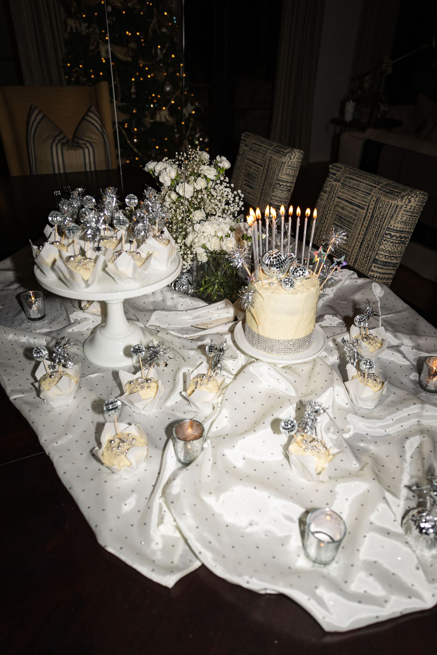Elegant birthday dessert table with lit cake, cupcakes, silver decorations, and white floral arrangement.