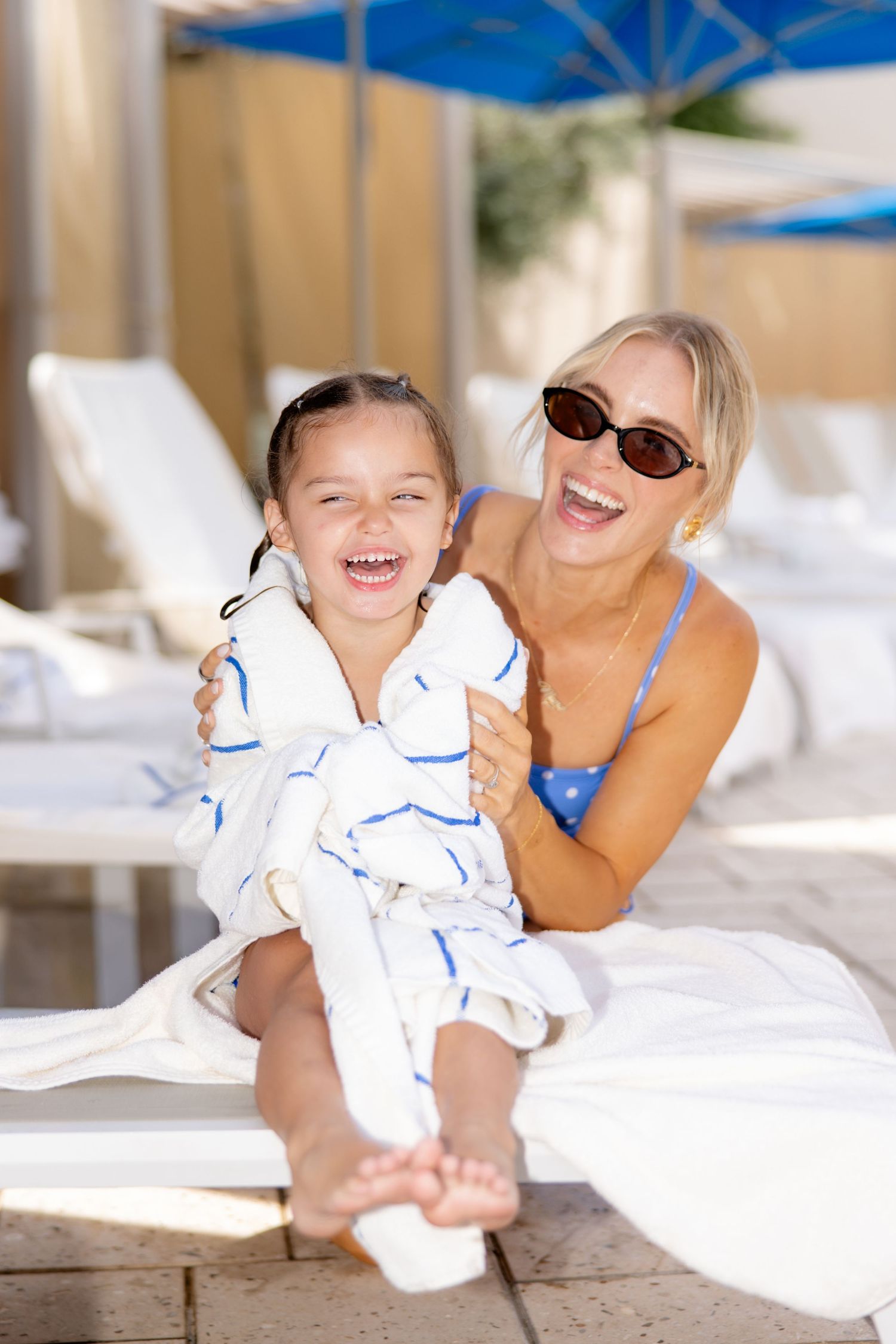 A laughing woman in a blue bikini and sunglasses hugs a giggling girl wrapped in a striped towel poolside.