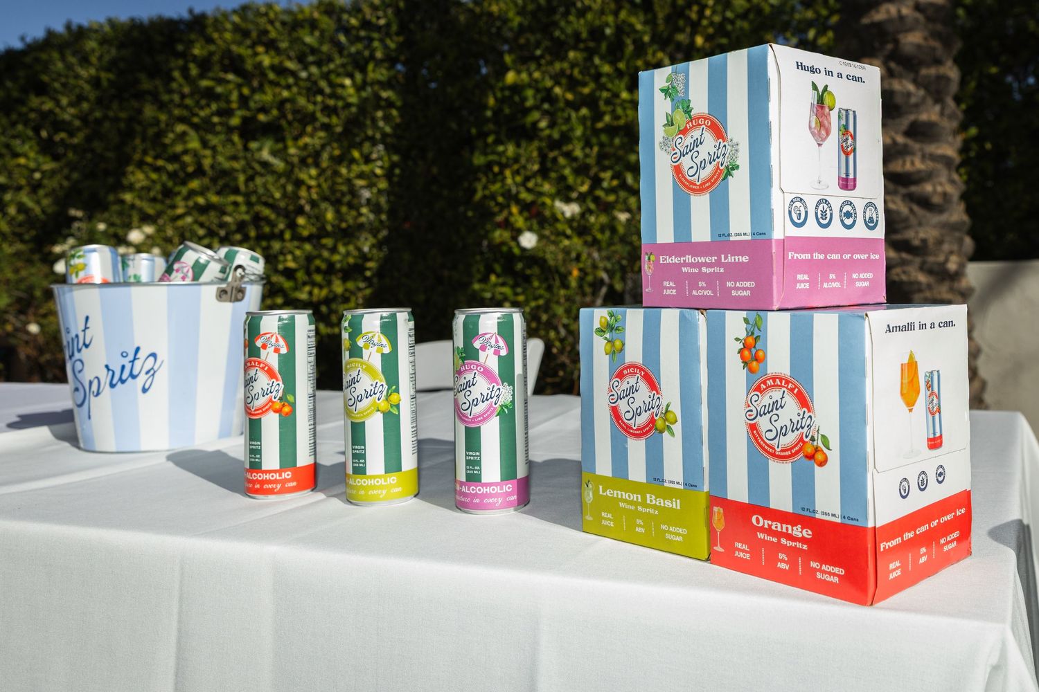Colorful cans and boxes of Saint Spritz non-alcoholic wine spritz displayed on a white table outdoors.