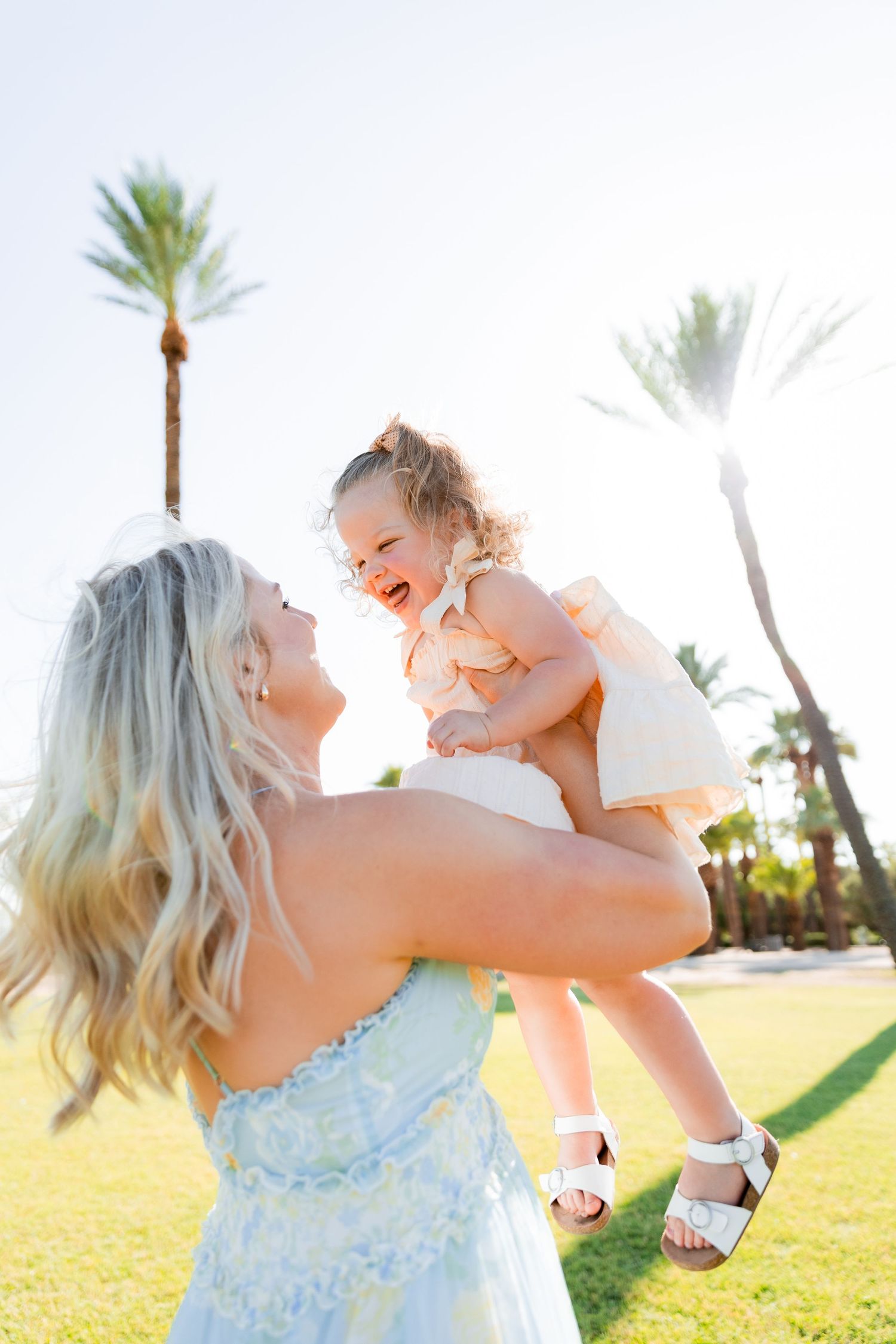 Mother lifting joyful toddler girl into the air in a sunny outdoor setting with palm trees.