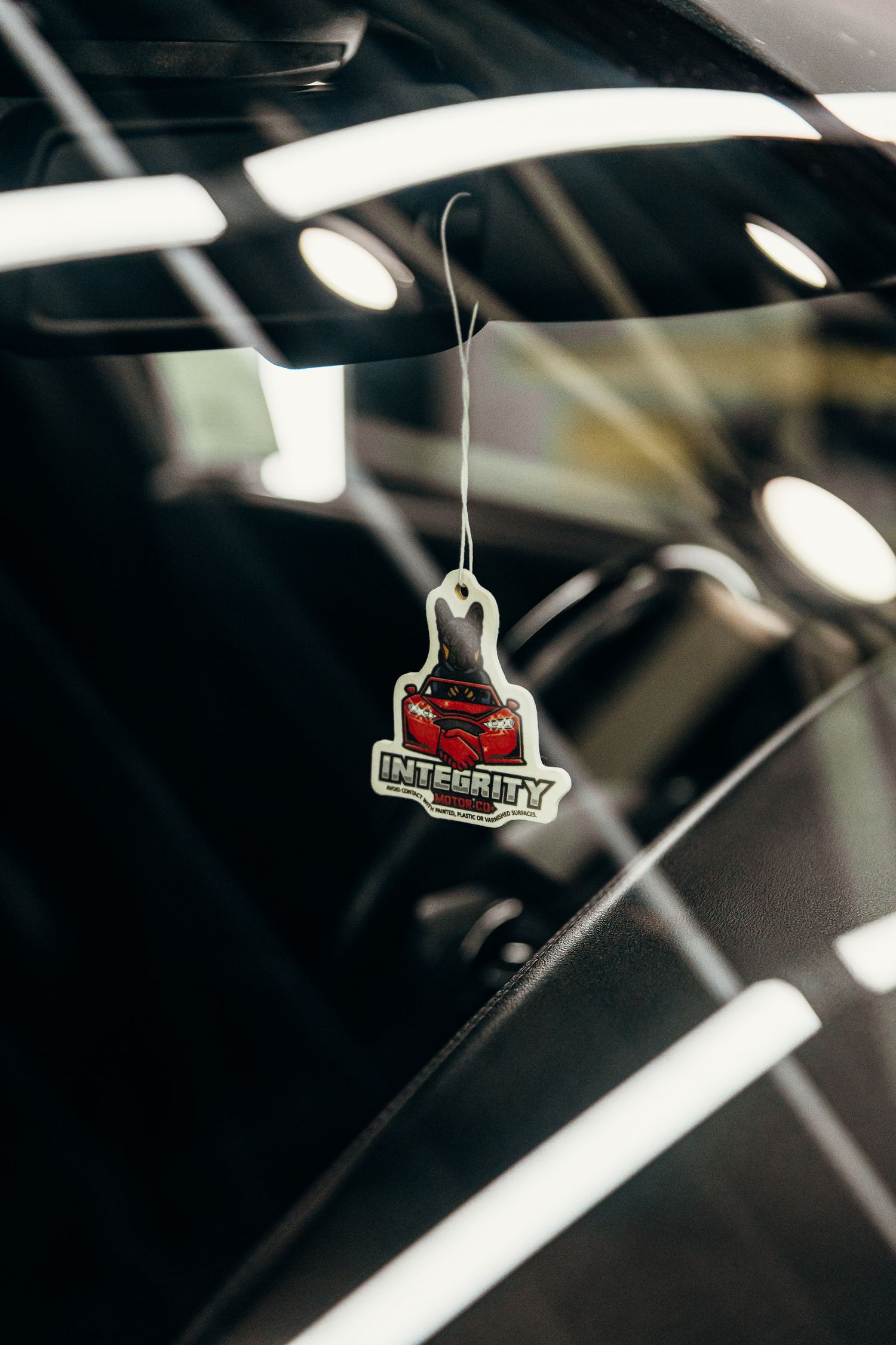 Integrity branded car air freshener hanging from rearview mirror featuring red sports car logo in dark vehicle interior.