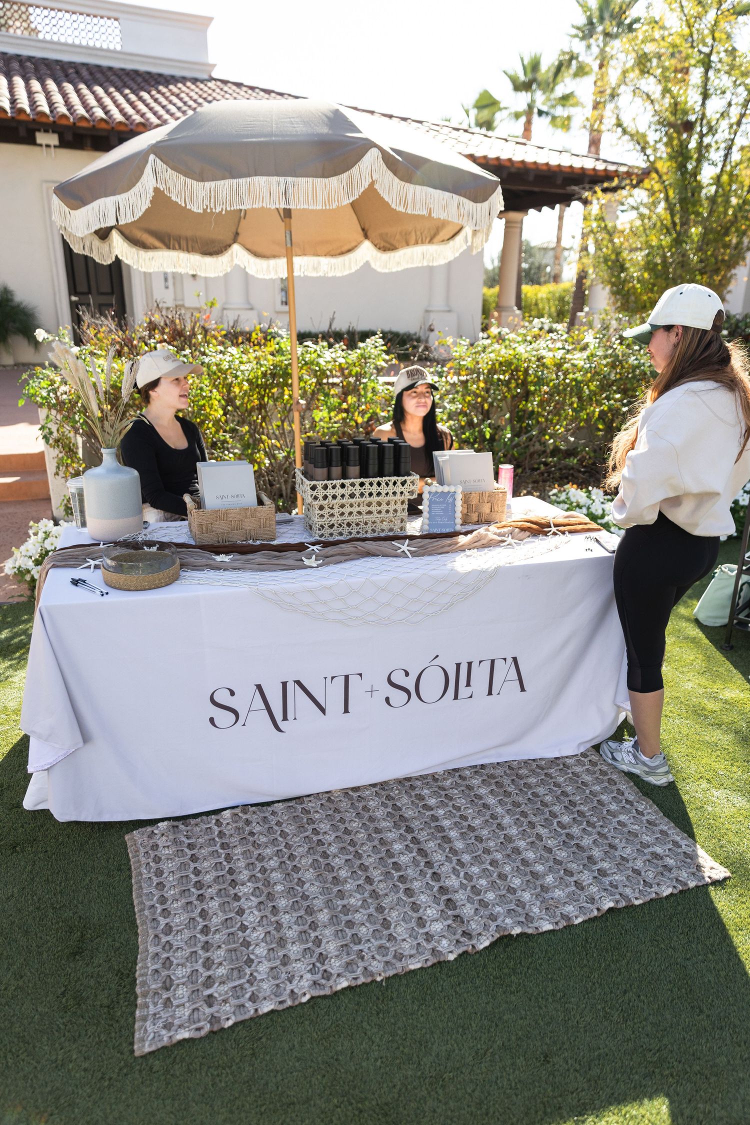 Two vendors at a Saint Solita branded booth with a fringed umbrella assist a customer at an outdoor market.