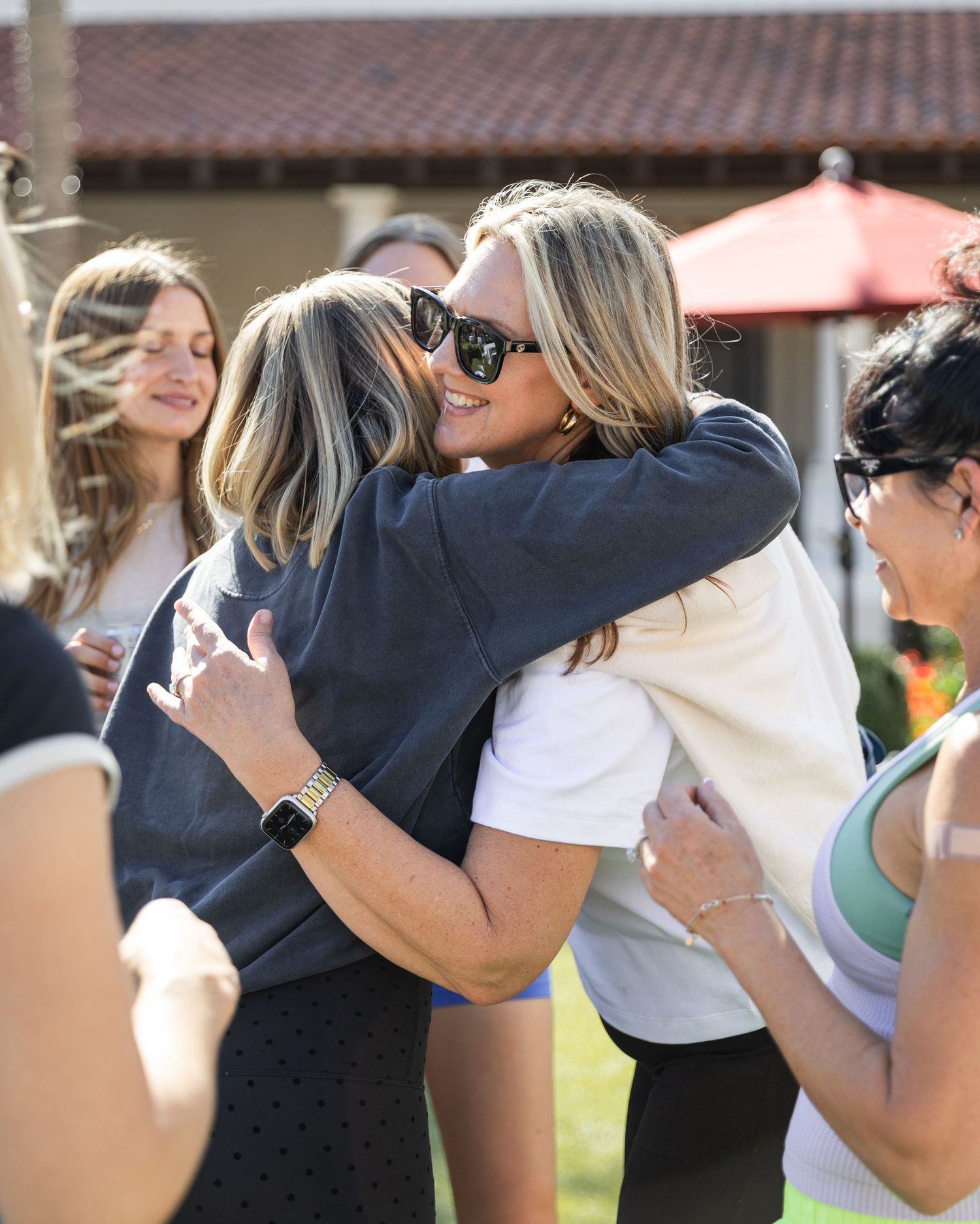 Two blonde women share a warm hug at an outdoor social gathering on a sunny day.