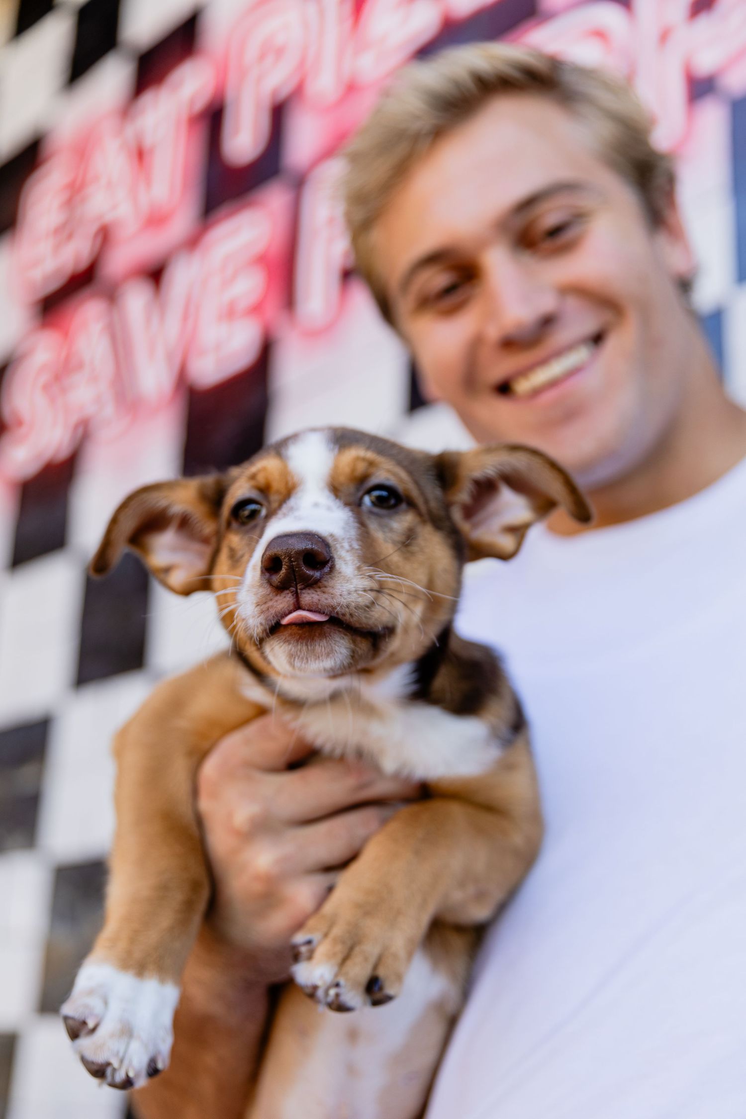 Smiling man holds adorable brown and white puppy in front of Eat Pizza Save Puppies neon sign.
