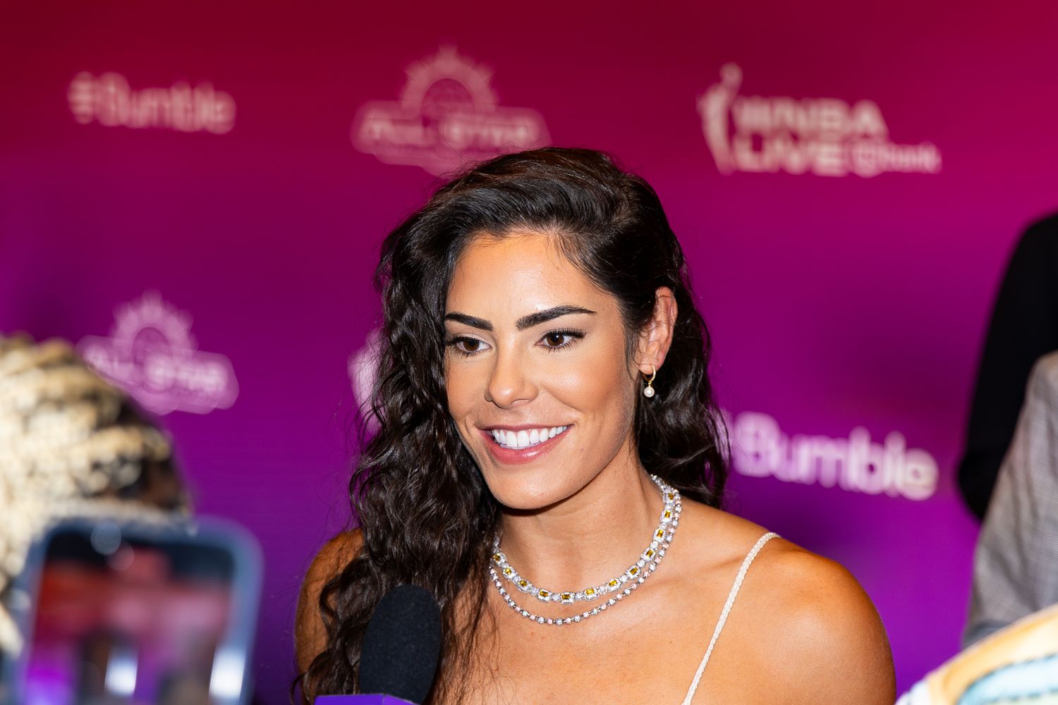 Smiling woman with dark curly hair wearing diamond necklace at WNBA All-Star Bumble event.