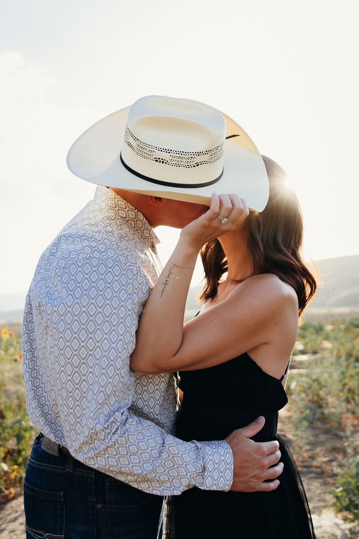 Couple sharing a romantic kiss outdoors, man wearing a white cowboy hat and patterned shirt, woman in black top.