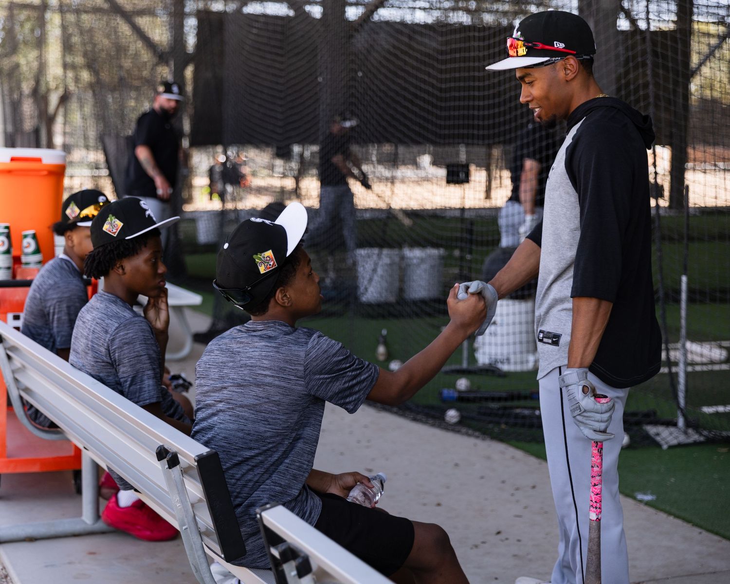 A baseball player talks to young players seated on a dugout bench during an outdoor practice session.