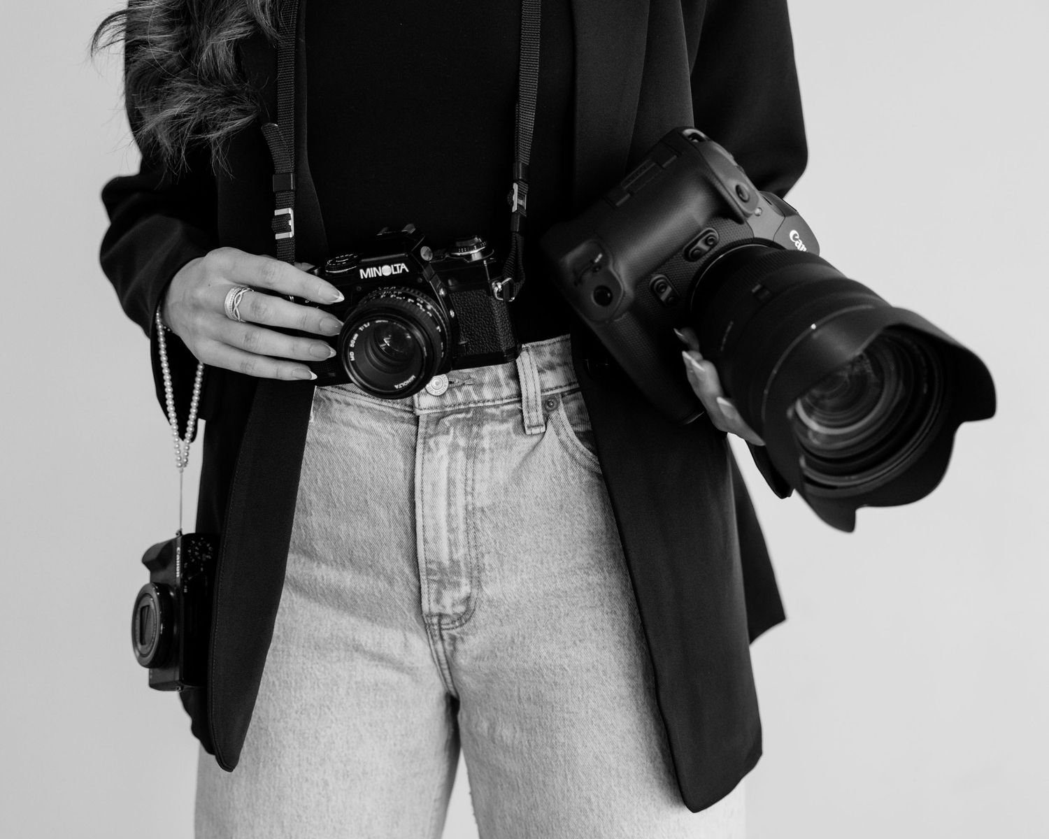 Black and white photo of a female photographer holding two cameras, wearing a black blazer and light jeans.