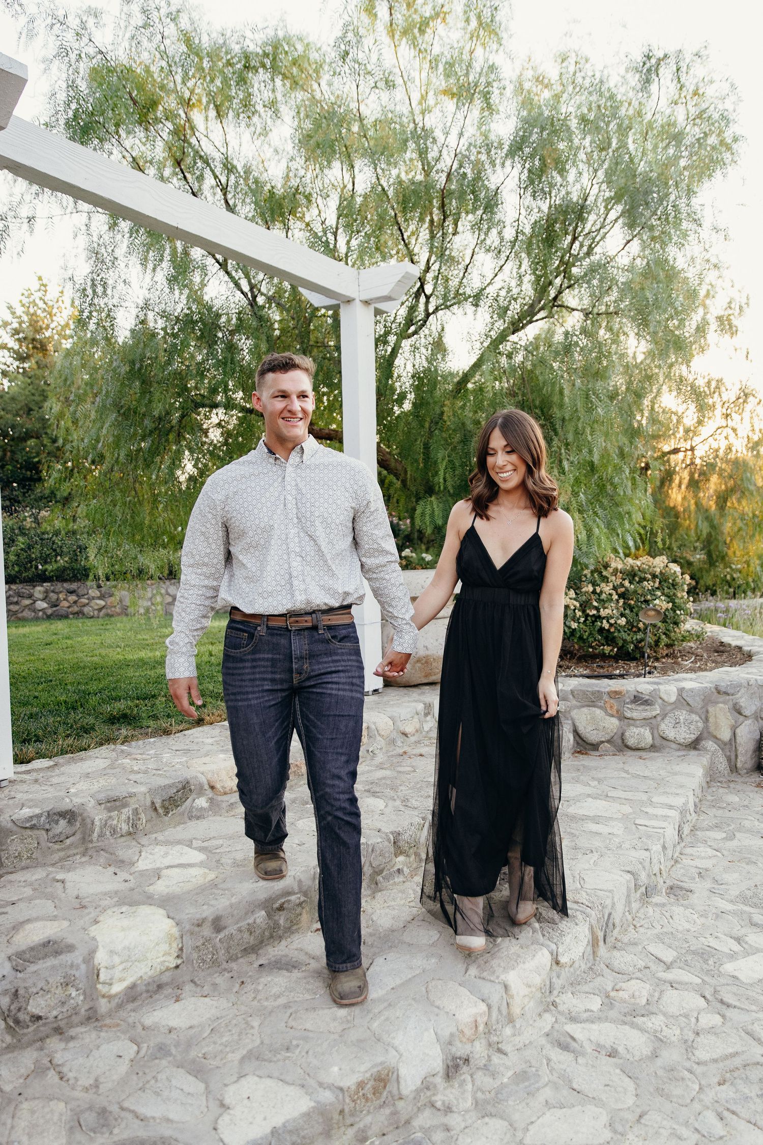 Couple holding hands walking down stone steps in a garden, man in jeans and shirt, woman in black maxi dress.