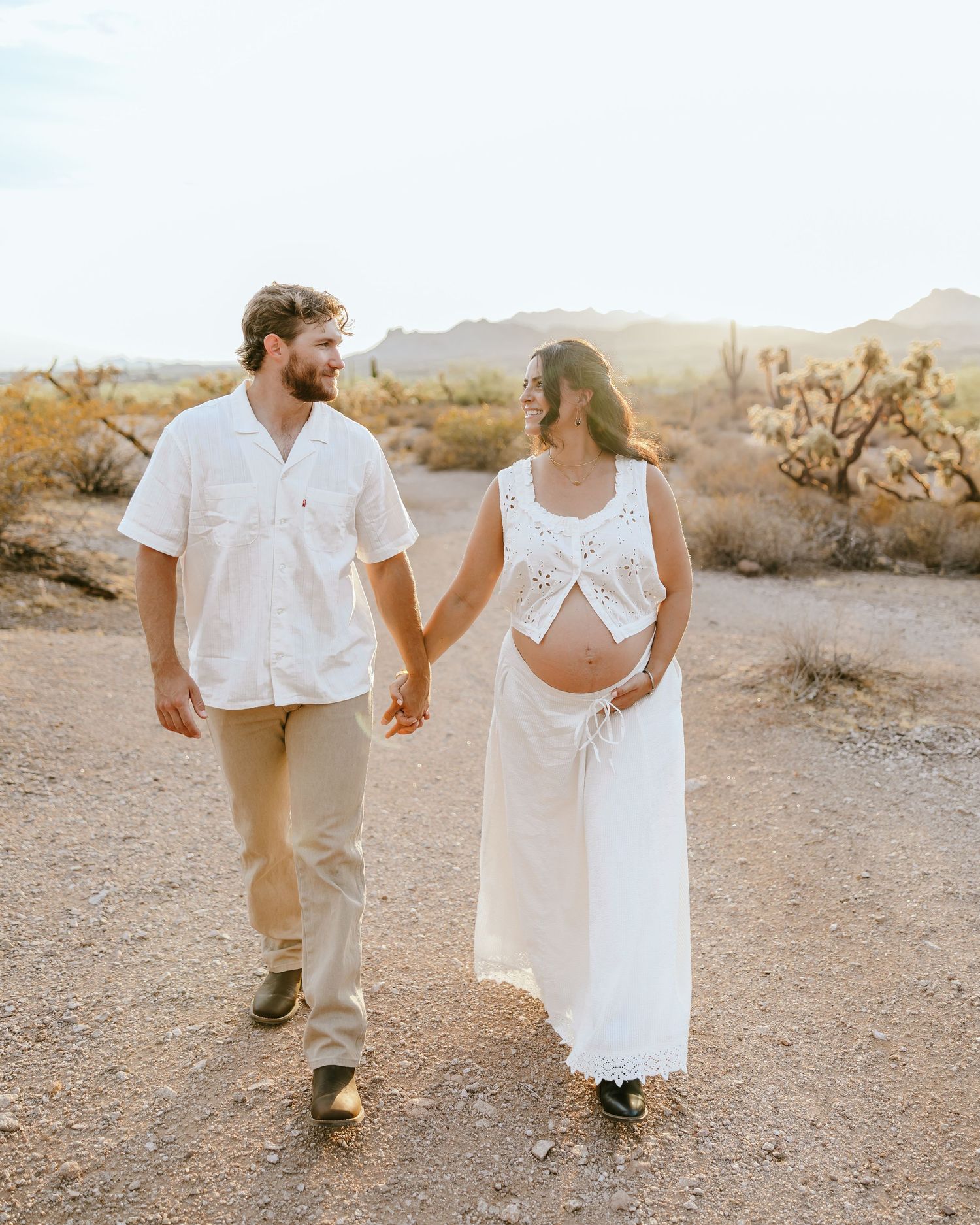 Couple in white outfits holding hands during desert maternity photoshoot at golden hour.