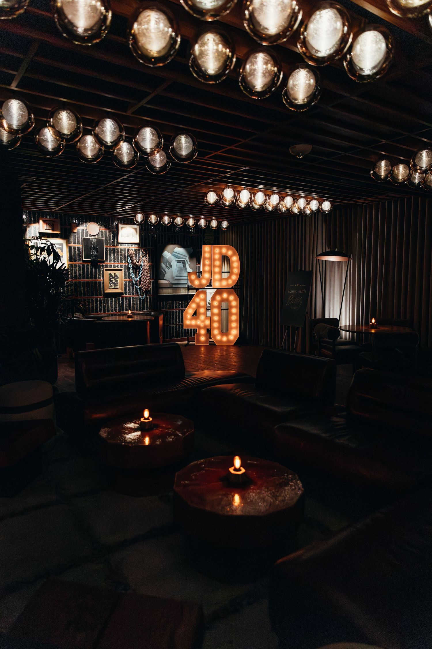 Elegant dark lounge venue with glowing JD40 marquee letters, pendant lights, and candlelit tables.