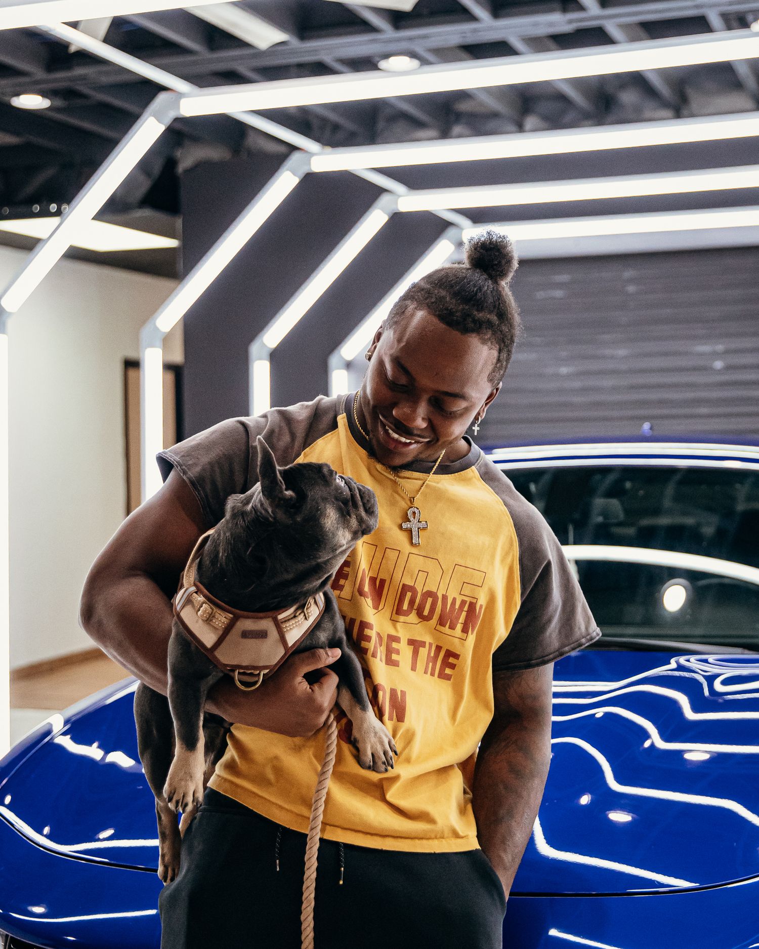 A muscular man with a bun hairstyle holds a French Bulldog in front of a blue sports car in a modern showroom.