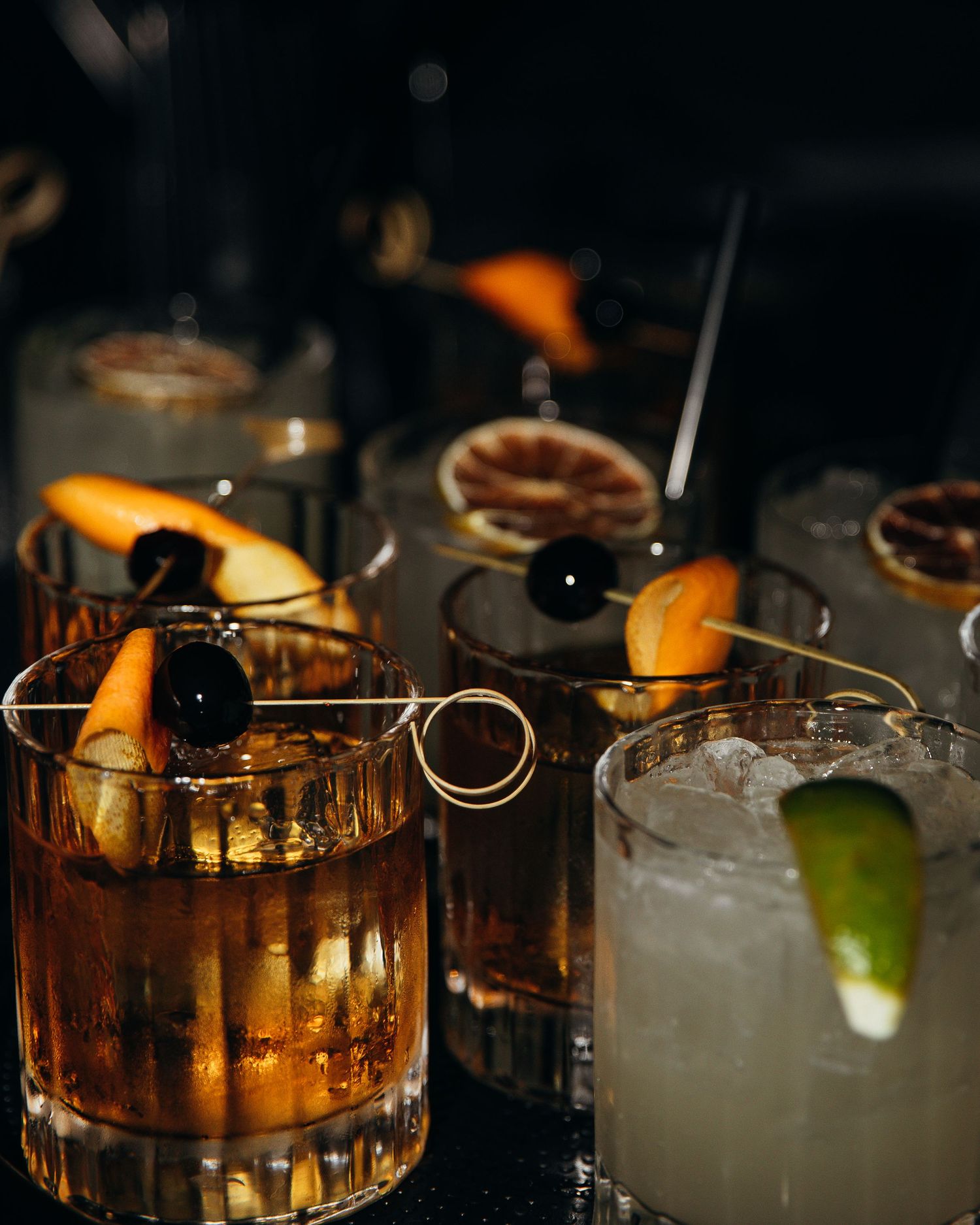 Craft cocktails garnished with orange slices and cherries served in rocks glasses at a bar.