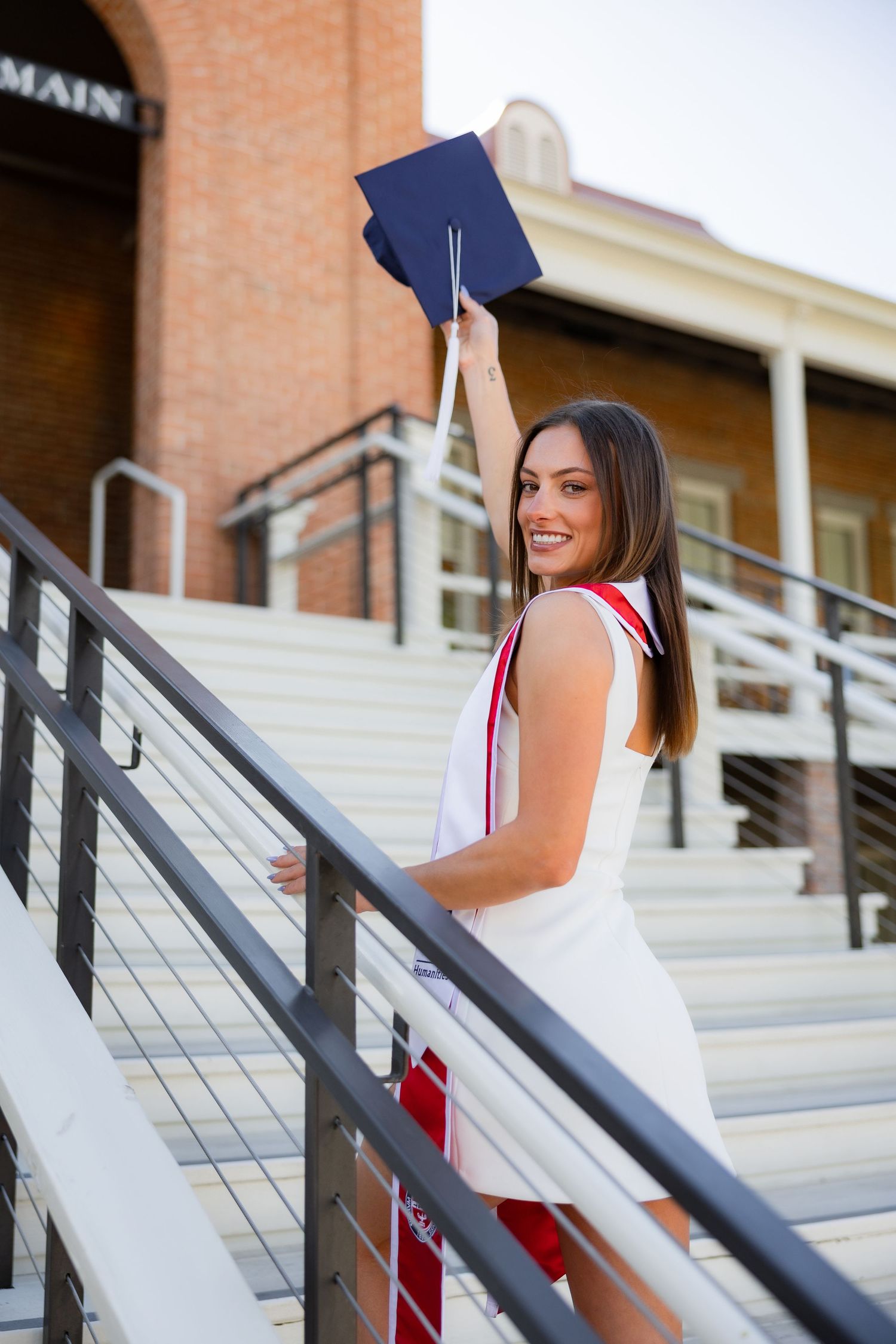 Smiling female graduate in white dress raises cap on stadium steps during graduation.