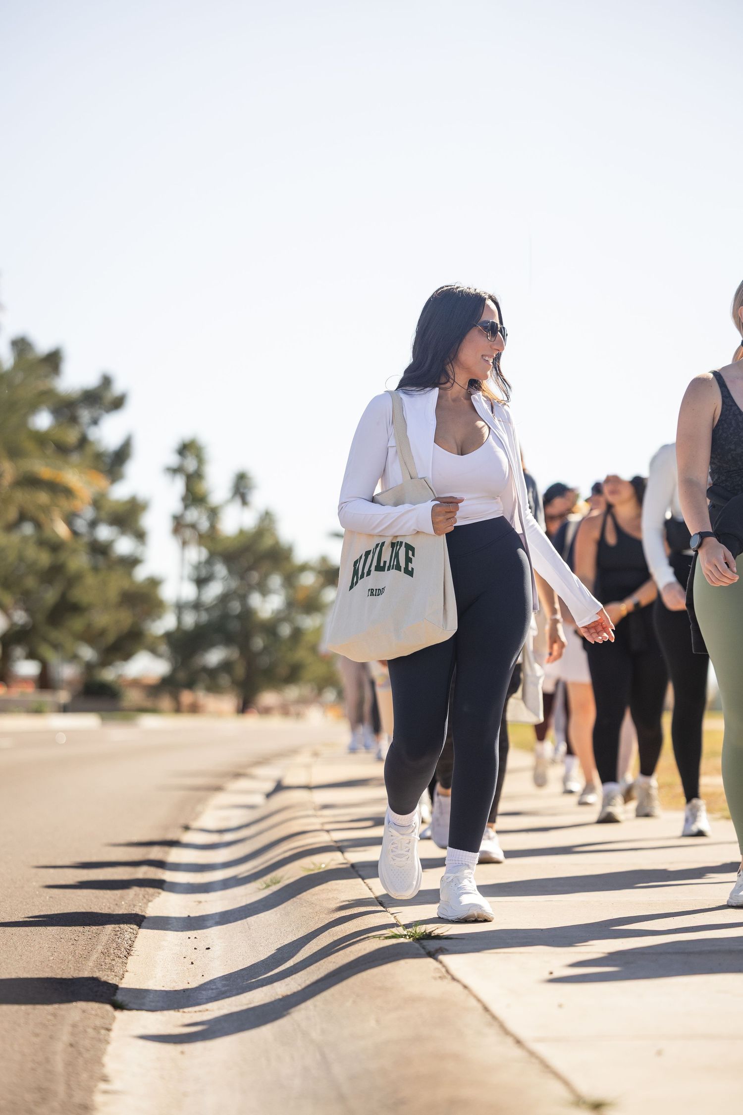 Woman in white top and black leggings carries a cream tote bag while walking with a group outdoors on a sunny day.