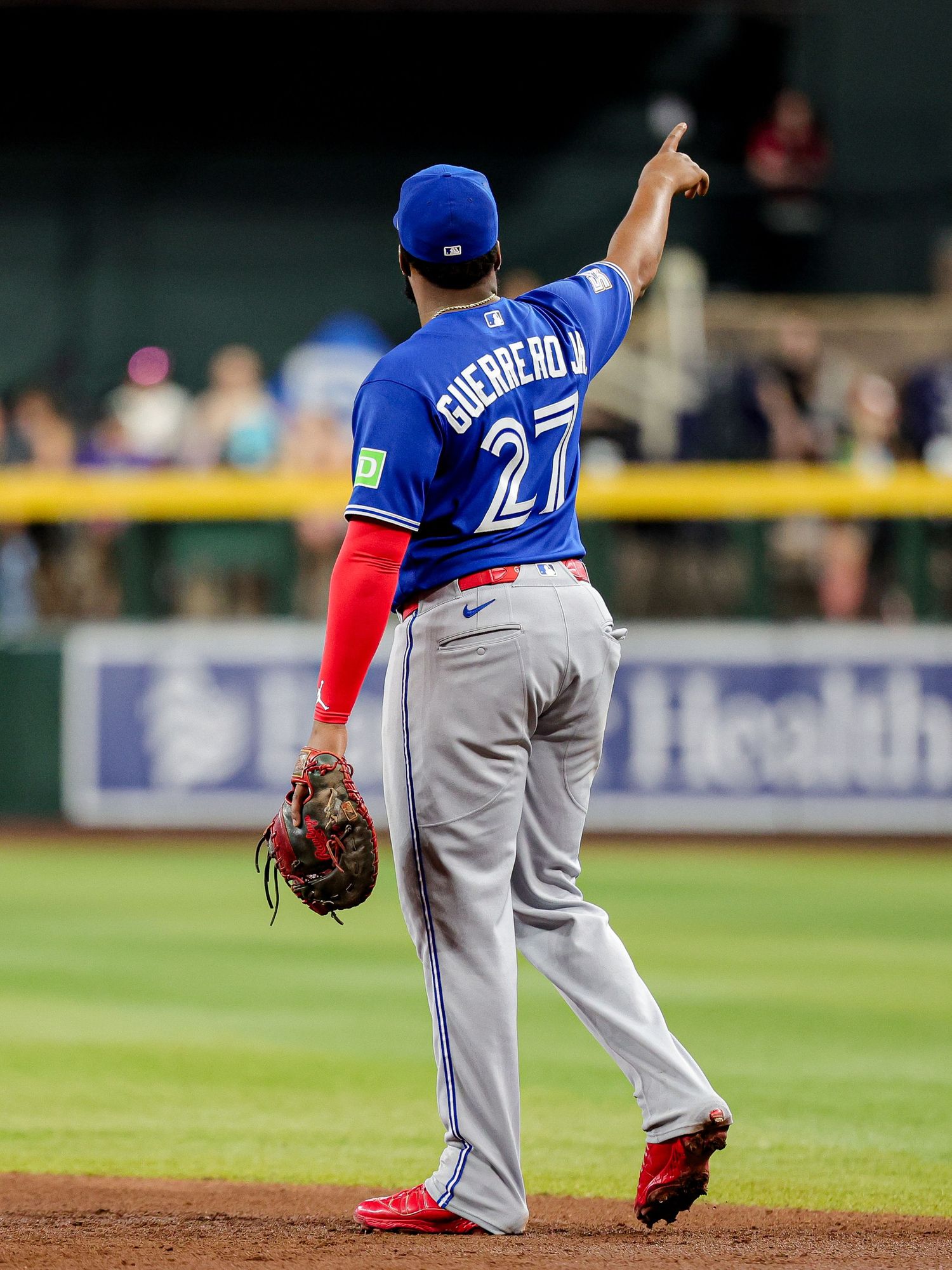 Toronto Blue Jays Guerrero Jr. #27 points upward while standing on the field in gray away uniform.