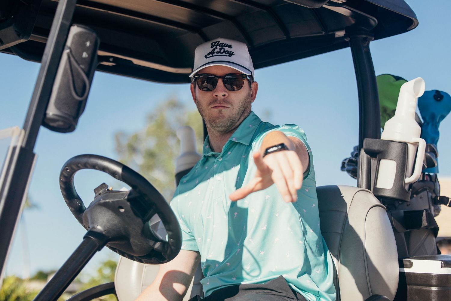 A man in a mint polo shirt and white cap points from a golf cart on a sunny day.