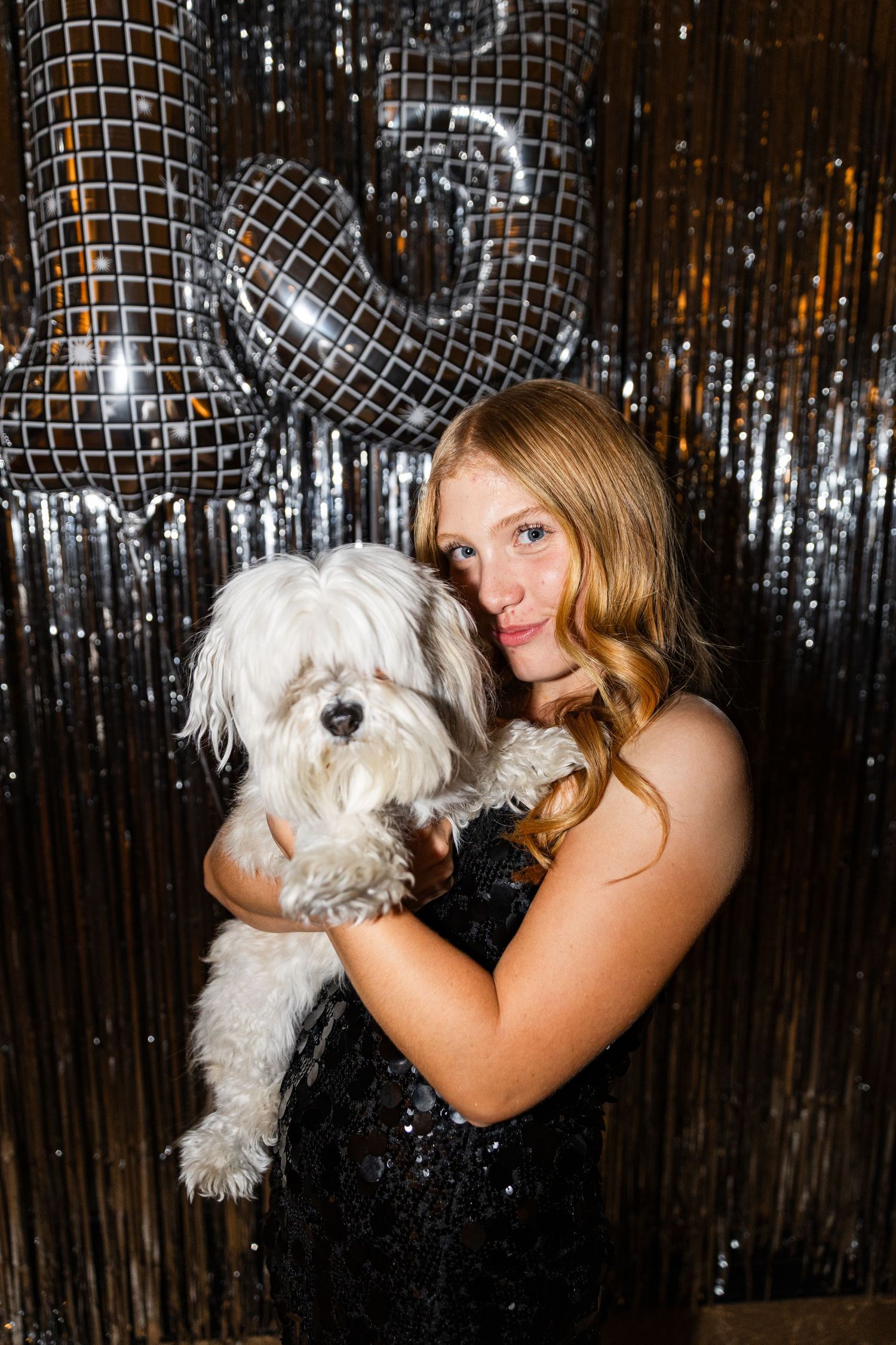 Teen girl in black sequin dress holding a fluffy white dog in front of silver disco balloon backdrop.
