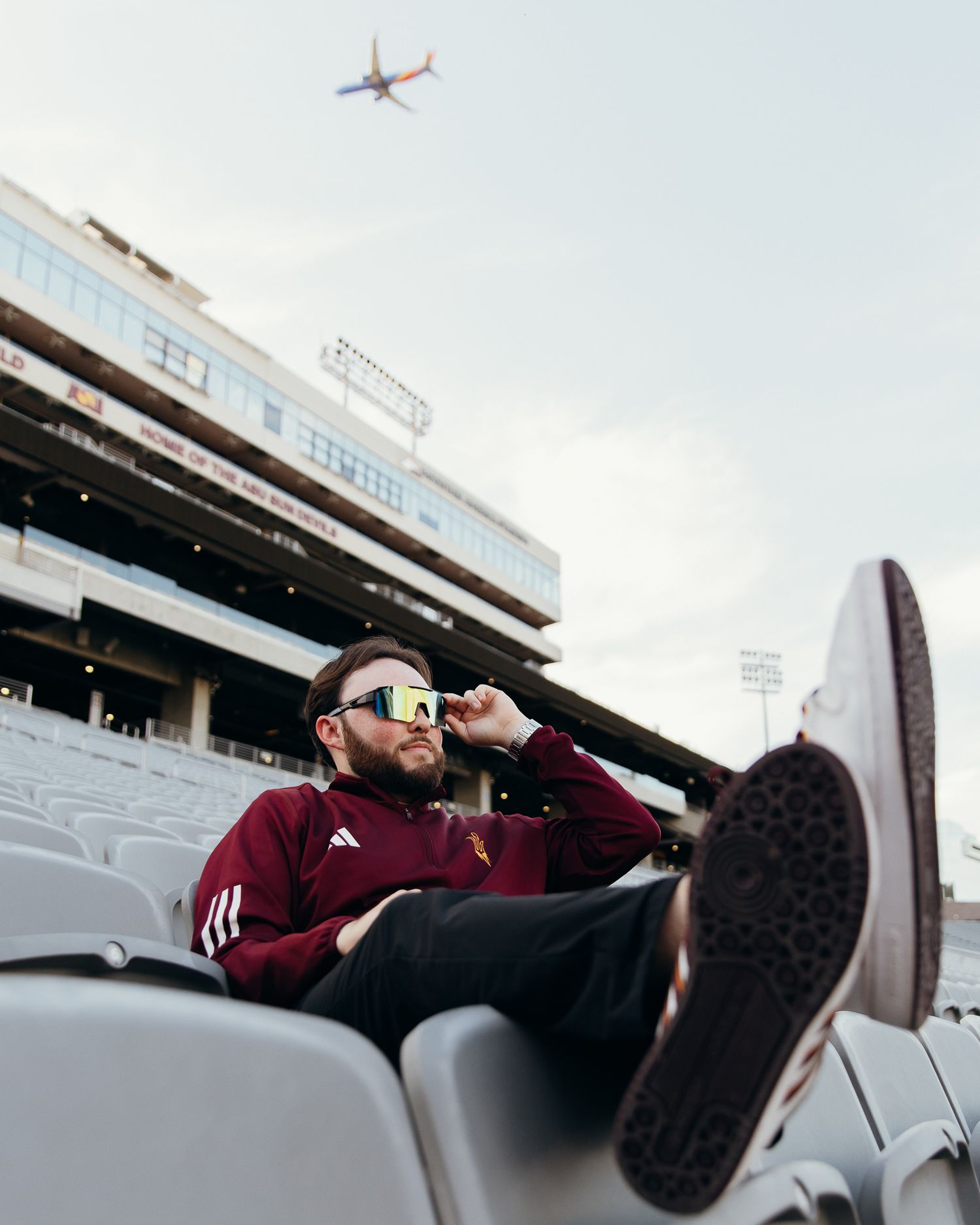 Young man in ASU maroon jacket wearing sunglasses relaxing in stadium seats.