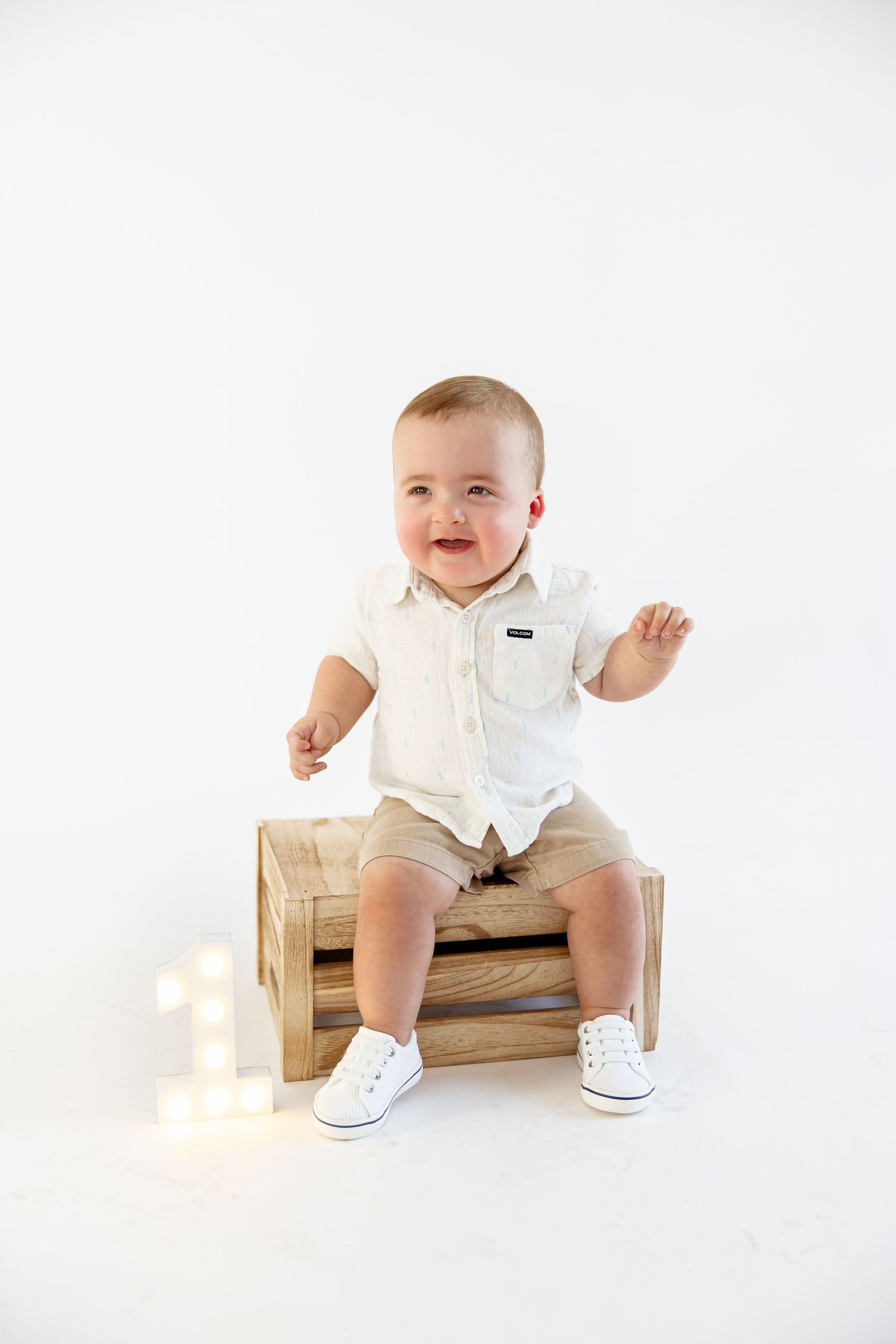 Adorable baby boy in white shirt sitting on wooden crate for first birthday photo session.