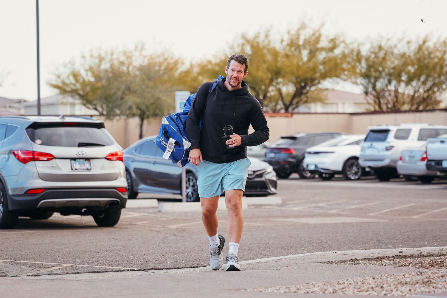 Man in casual clothes walking through a sunny parking lot carrying a blue sports bag.