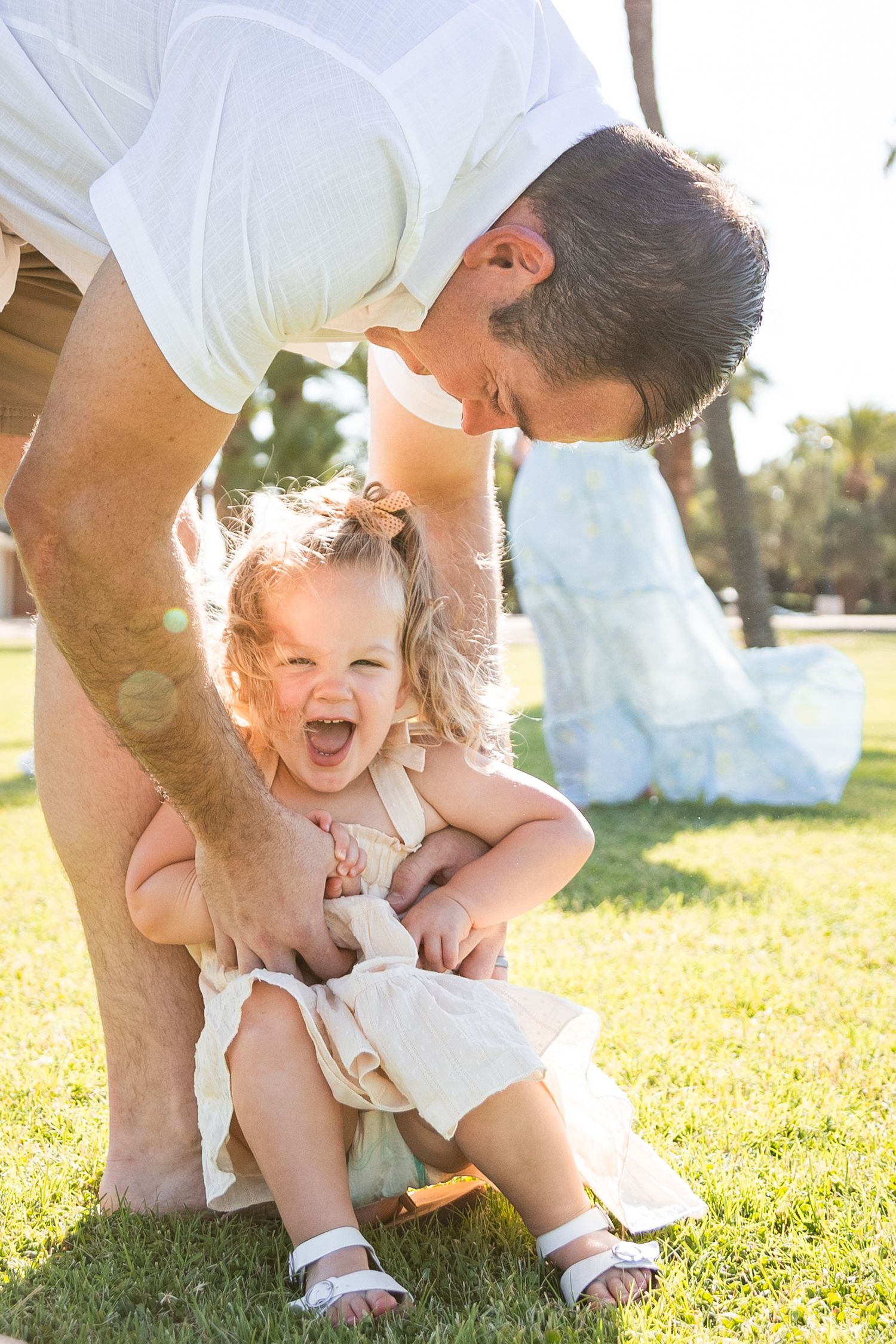 Father playfully lifting laughing toddler girl on a sunny green lawn with palm trees.