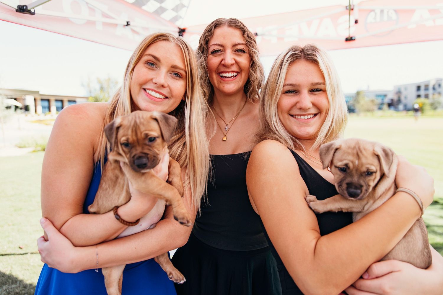 Three smiling women holding adorable puppies at an outdoor charity golf event under a tent.