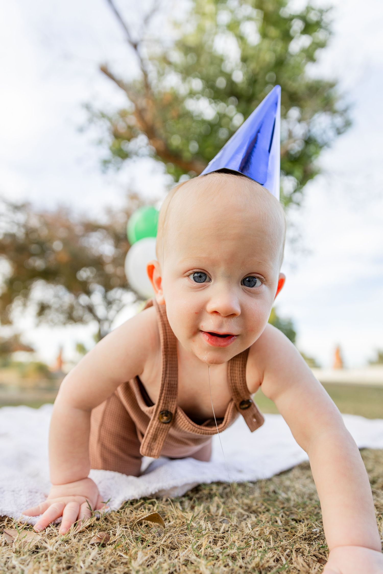 A curious baby in brown overalls and a blue party hat crawls on grass surrounded by balloons.
