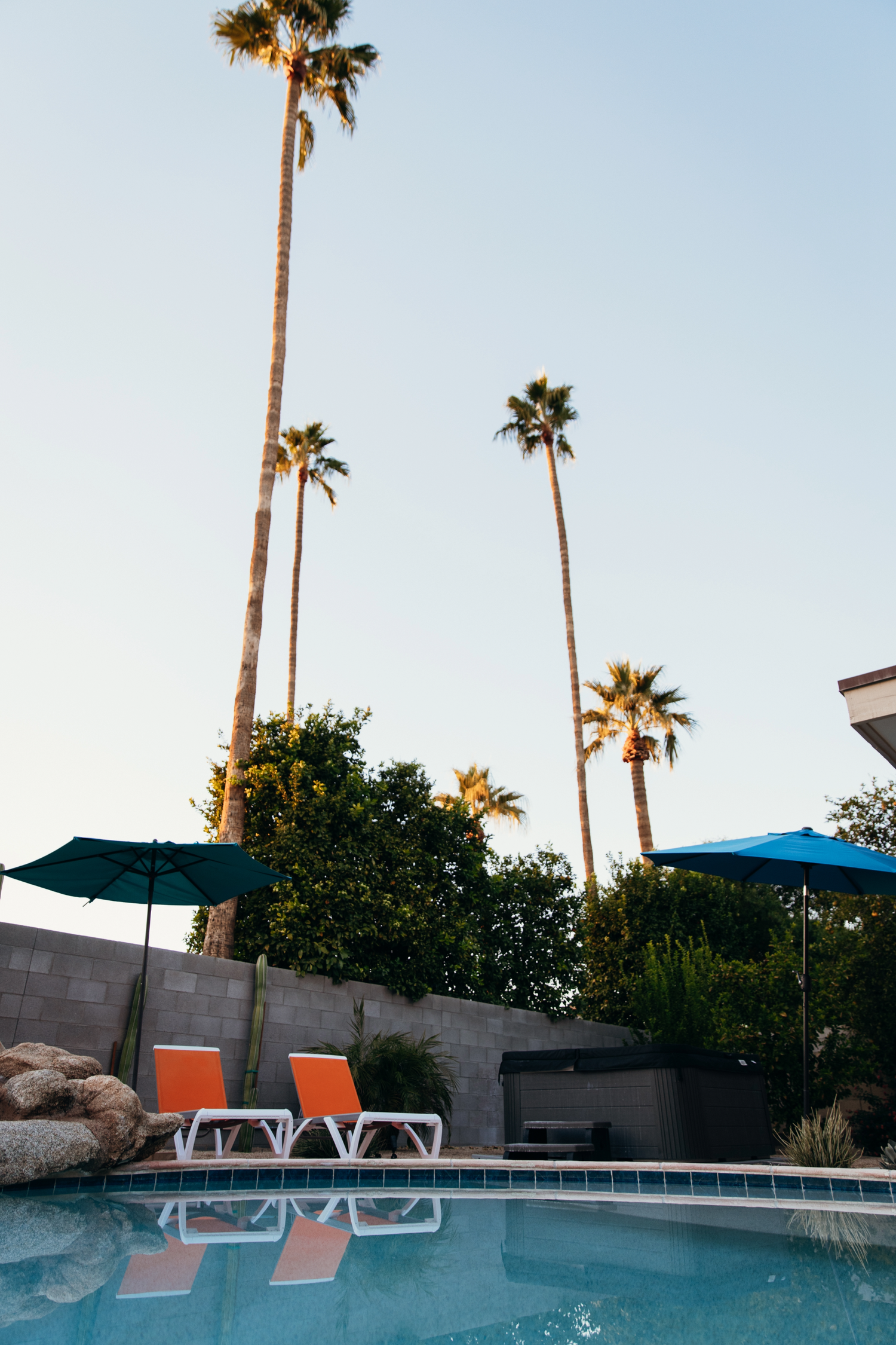 Orange lounge chairs beside a pool with teal umbrellas and tall palm trees at dusk in Palm Springs.