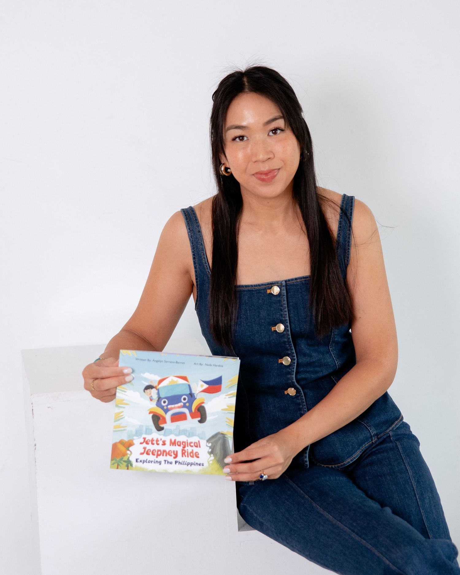 Woman in denim jumpsuit holding a children's book titled Jeff's Magical Jeepney Ride Exploring The Philippines.