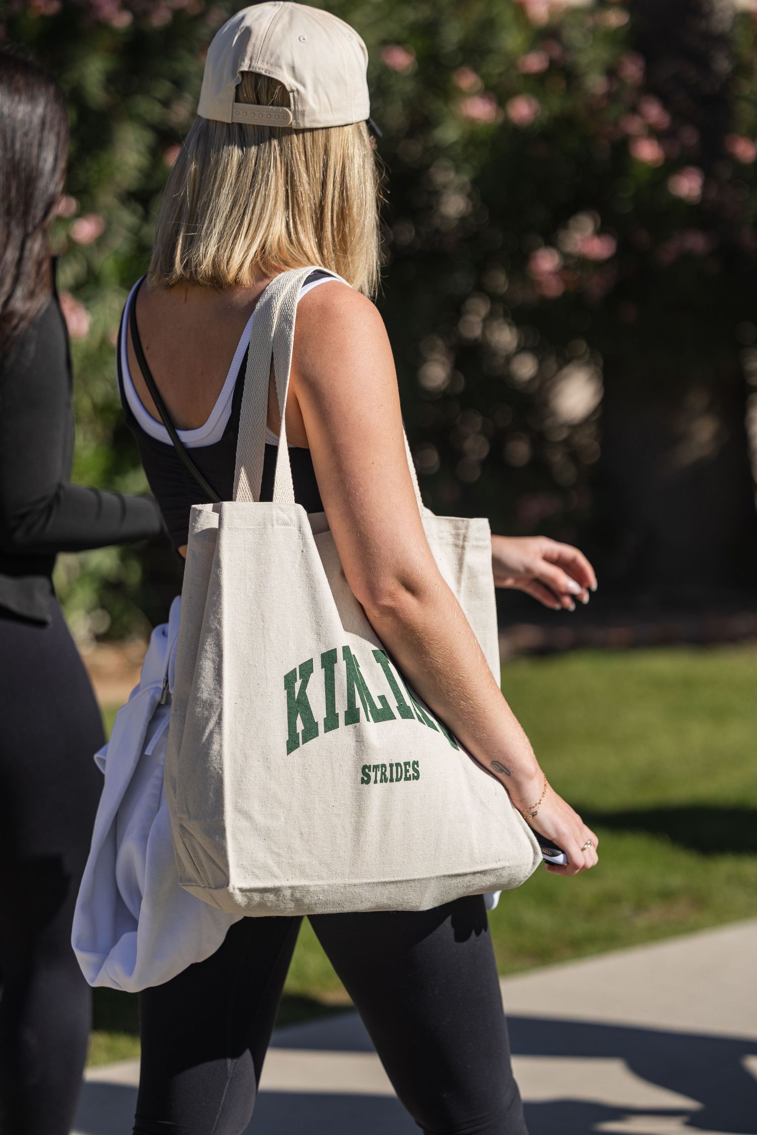 Blonde woman wearing a baseball cap and black leggings carries a cream tote bag with green lettering outdoors.