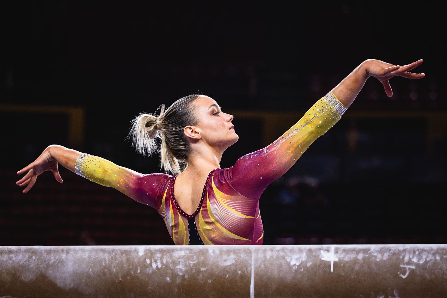 ASU gymnast extends arms gracefully on the balance beam in a vibrant yellow and pink leotard.