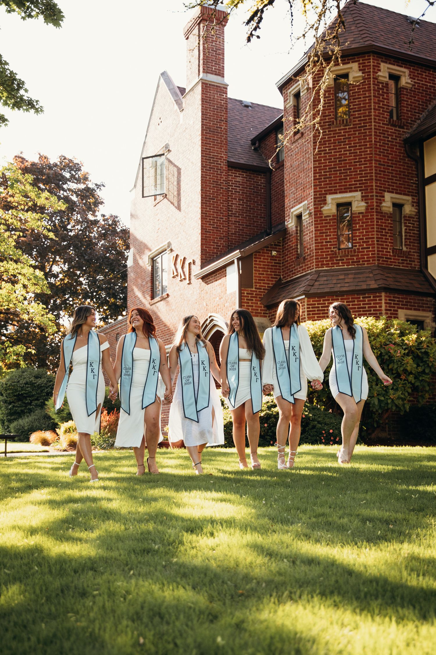 Group of female graduates in light blue stoles walking on lawn near brick house.