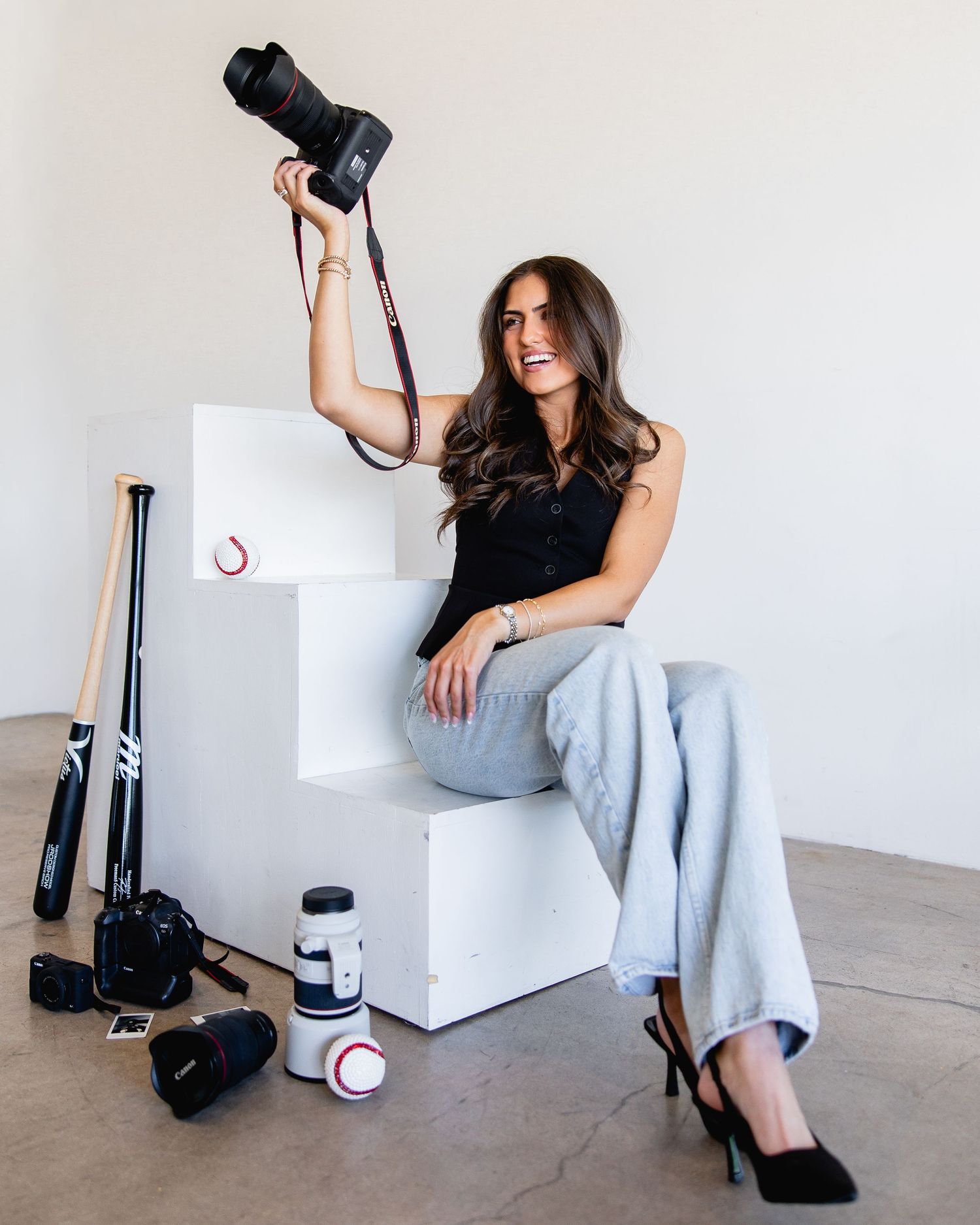 A woman photographer poses on white steps holding a Canon camera, surrounded by sports equipment and photography gear.