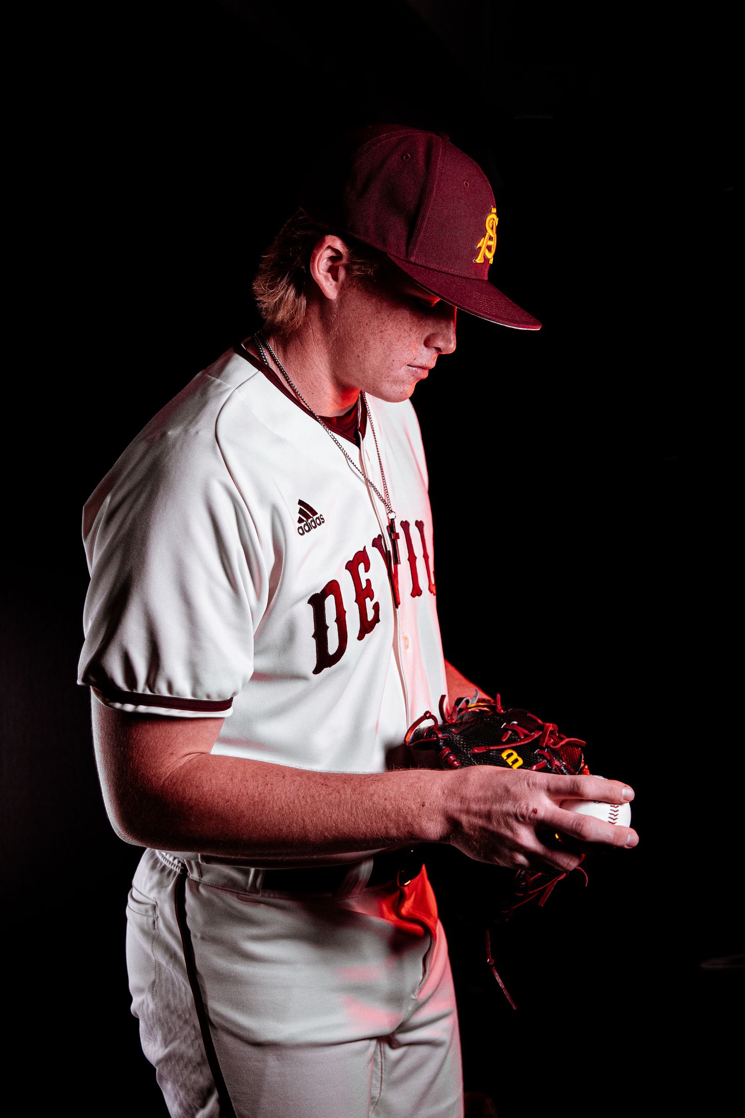 Arizona State Sun Devils pitcher examines baseball while wearing white Devils Adidas uniform and maroon cap.
