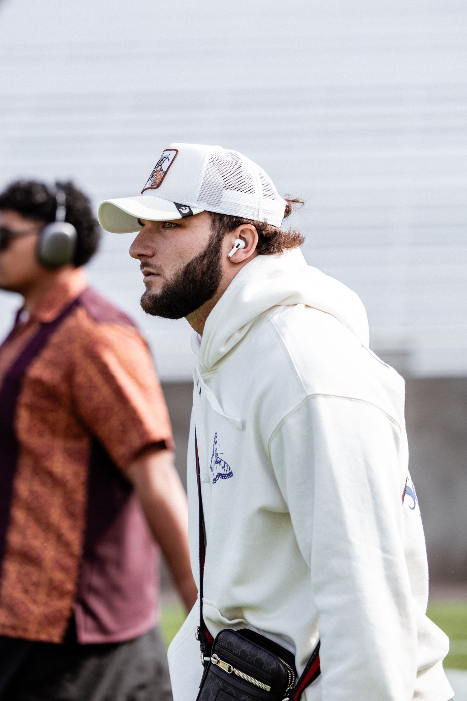 Young man in white hoodie and cap with AirPods walks at an ASU football event.