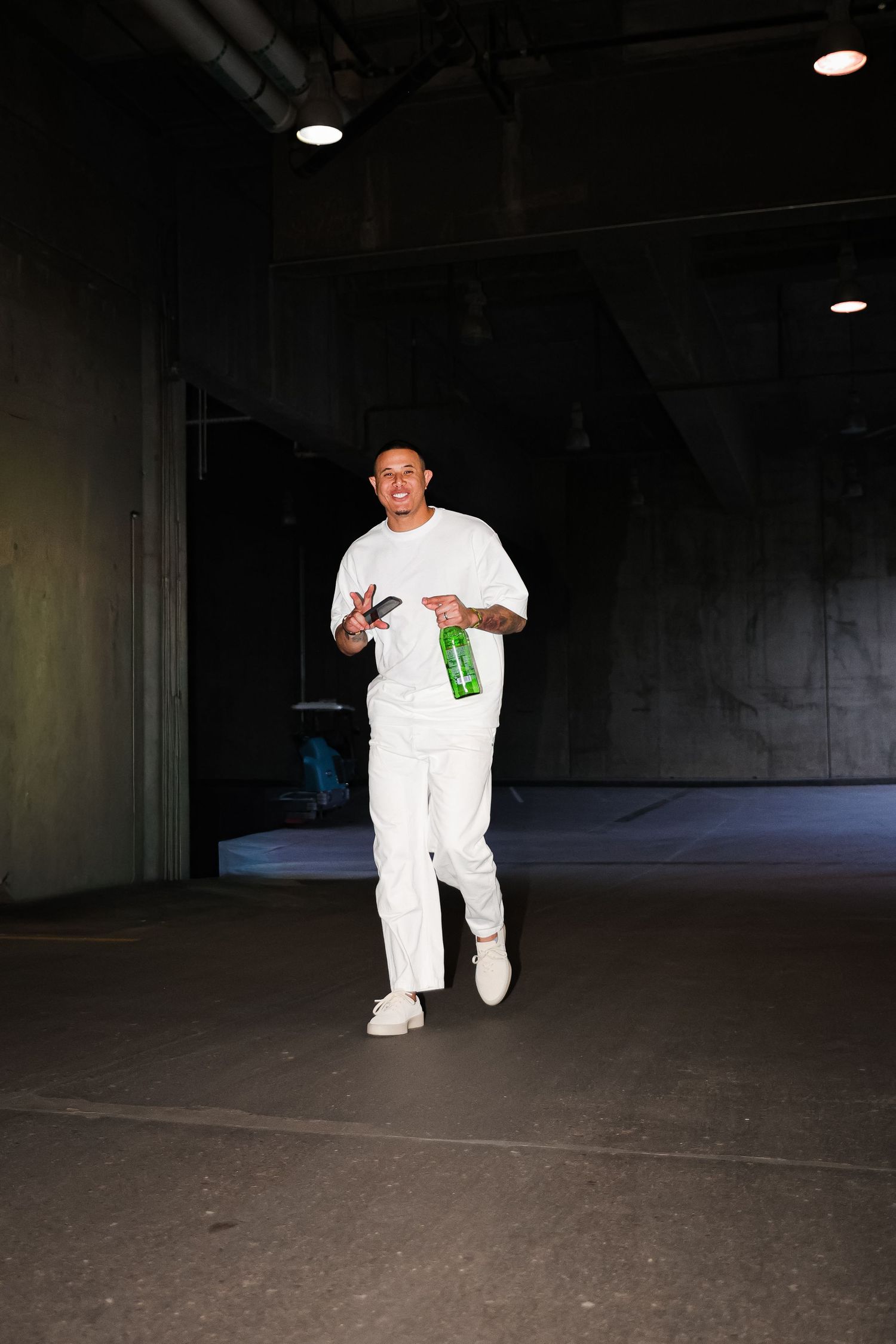 A young man in all-white outfit walks confidently through a dark tunnel holding a green bottle.