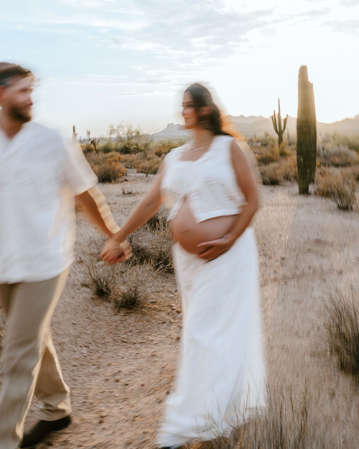 Pregnant woman in white dress holds hands with partner in desert landscape with saguaro cacti at golden hour.
