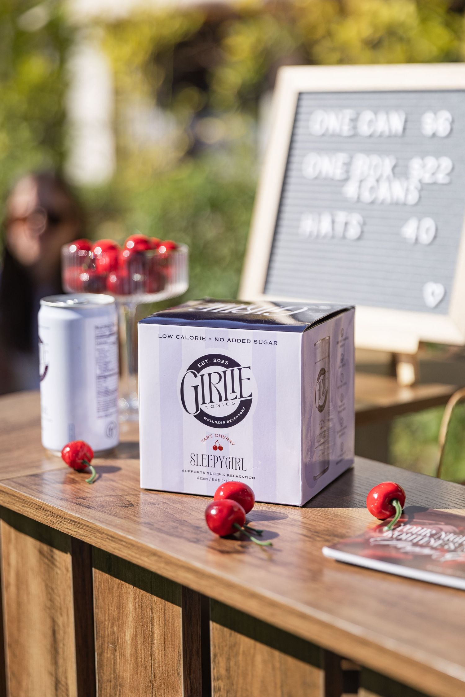 Girlie brand Sleepy Girl canned beverage box displayed on wooden table with fresh cherries and pricing sign.
