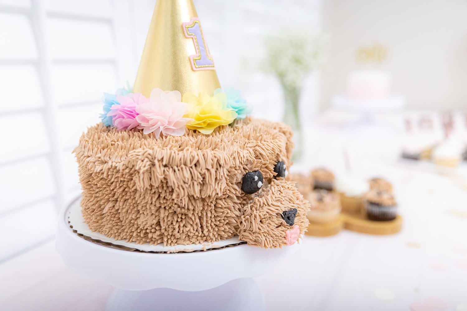 Adorable dog-shaped birthday cake with gold party hat and colorful pompoms on display.