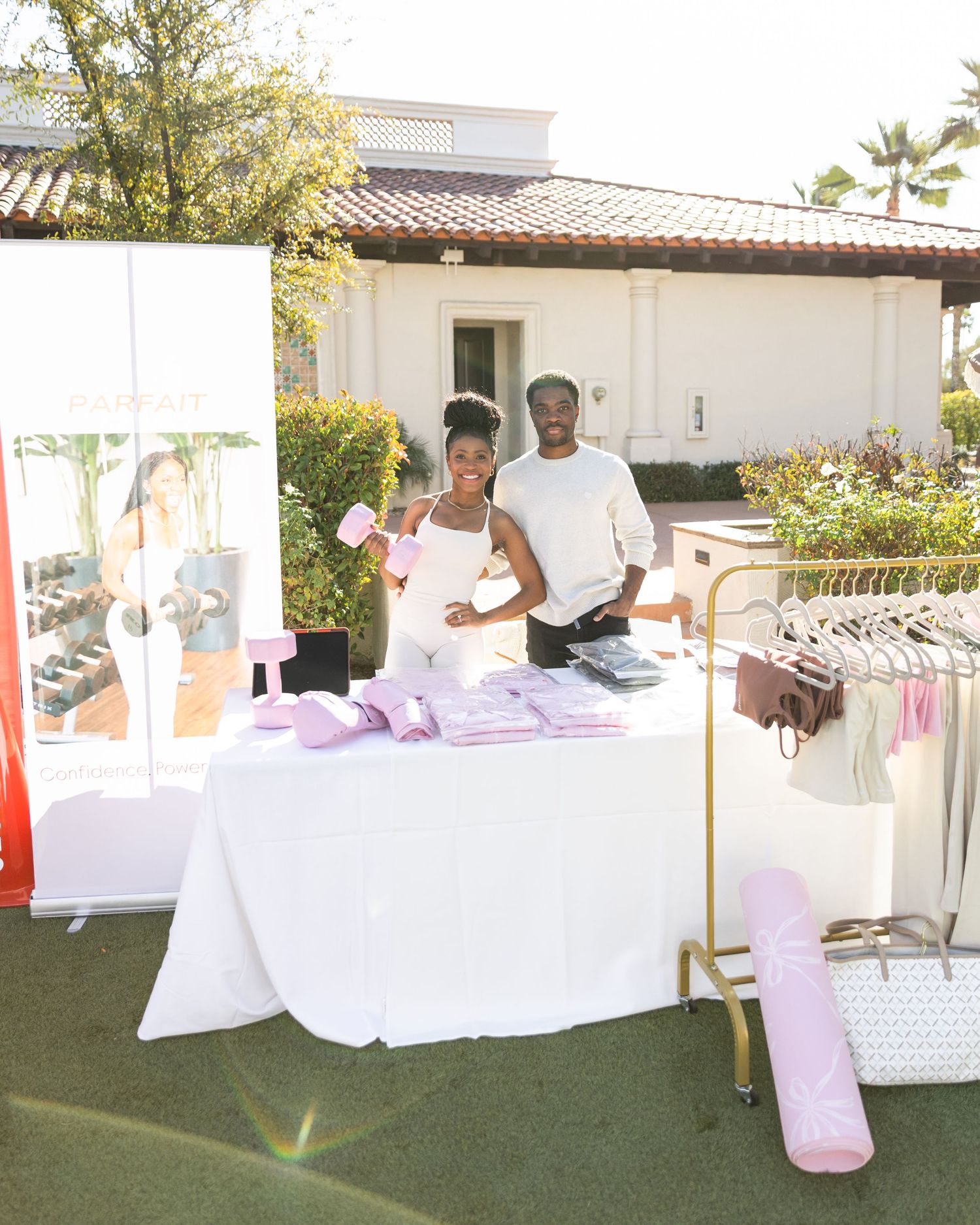 Two vendors smile behind a white table displaying pink activewear products at an outdoor pop-up market event.